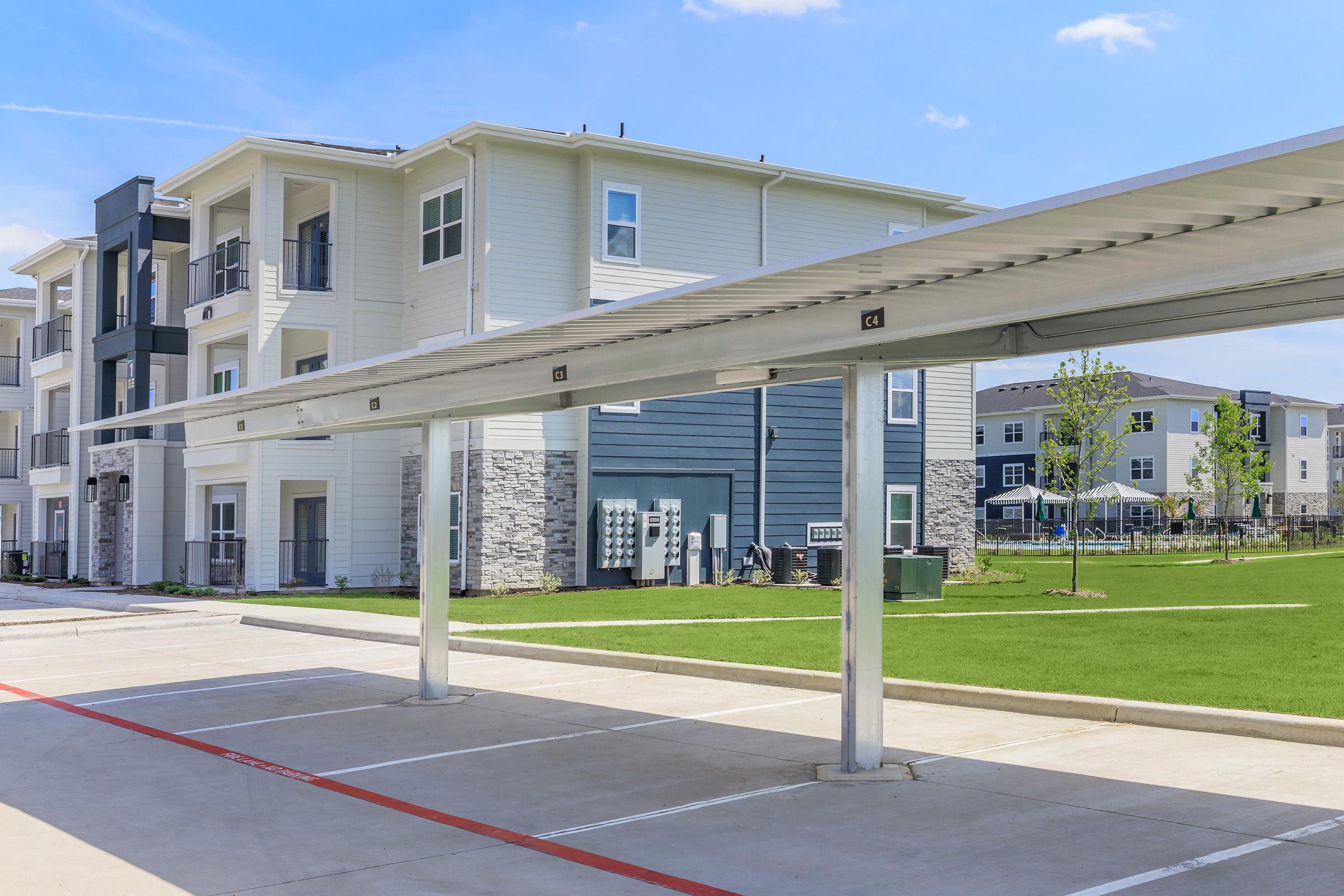 Modern apartment complex with multiple buildings featuring a mix of light and dark siding. In the foreground, there are covered parking spaces with a neatly maintained green lawn. The sky is clear with a few clouds, contributing to a bright, inviting atmosphere.