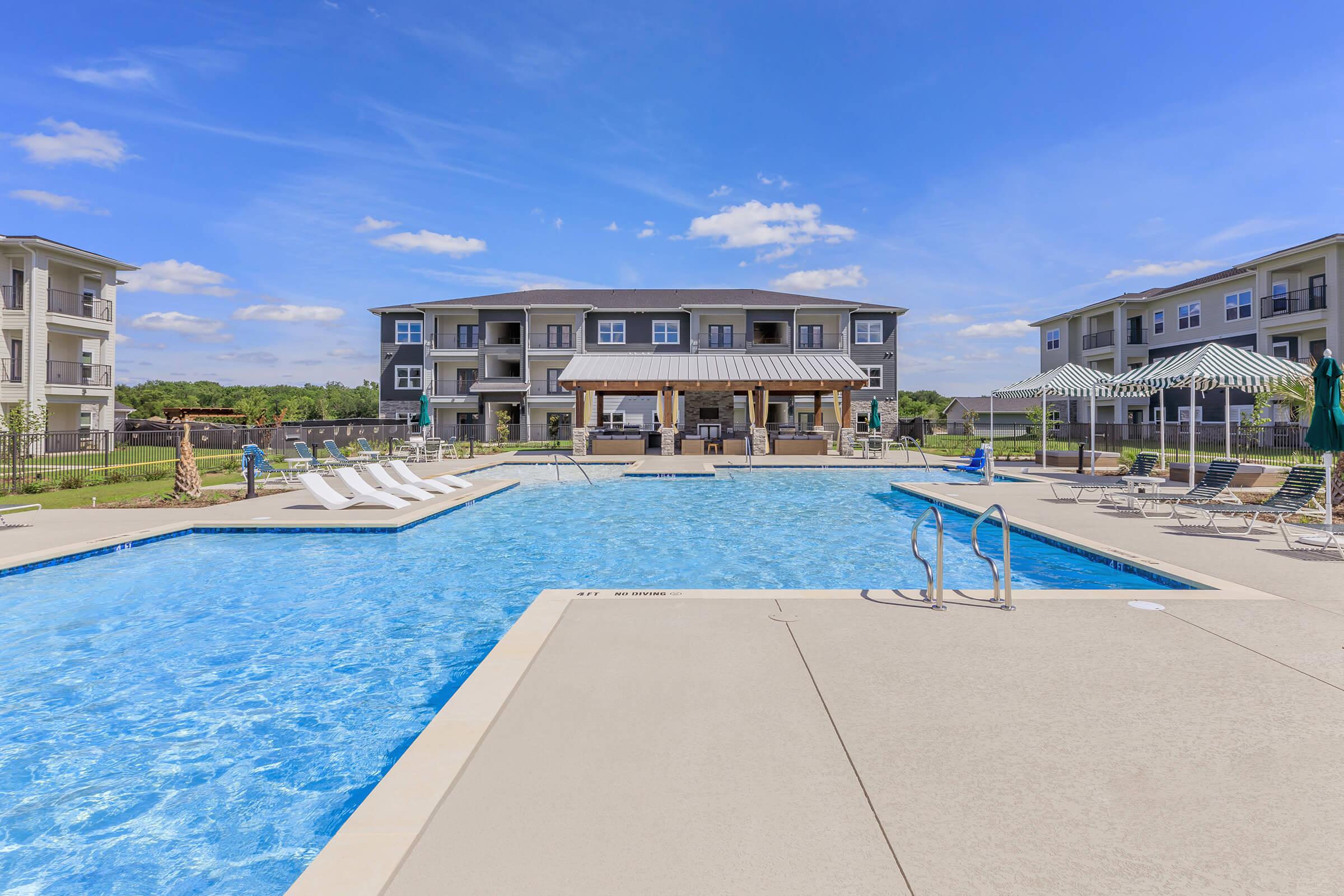 A bright outdoor swimming pool with lounge chairs and shaded areas, surrounded by modern apartment buildings. The clear blue water reflects the sunny sky with few clouds, creating a relaxing atmosphere for residents and visitors.
