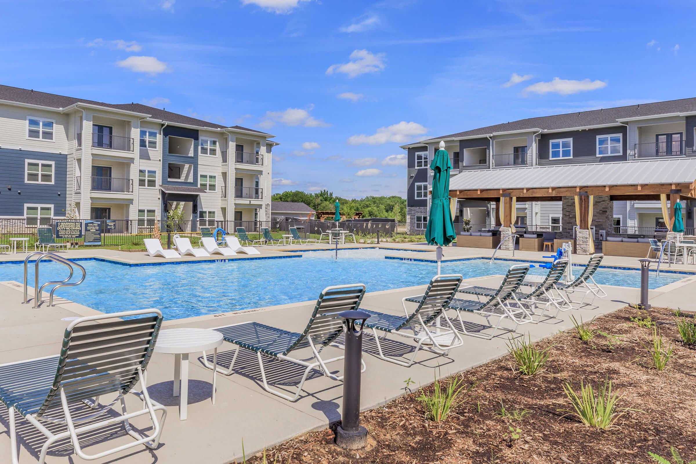 A bright outdoor swimming pool surrounded by lounge chairs, situated between two modern apartment buildings. There are green umbrellas and a shaded area with seating nearby. The scene is set against a clear blue sky with a few clouds, creating a relaxed and inviting atmosphere.
