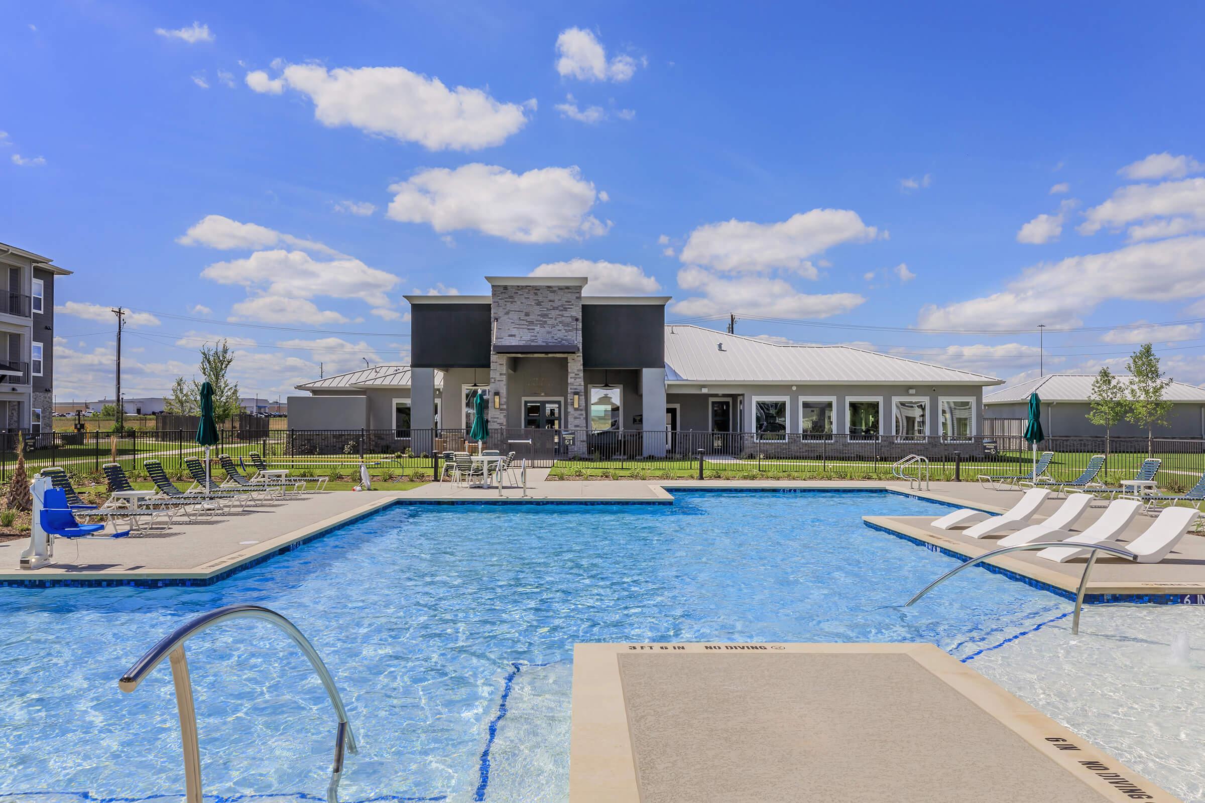 A clear blue swimming pool with lounge chairs around it, set against a backdrop of a modern building and a bright blue sky with scattered clouds. Green grass and trees surround the pool area, creating a welcoming outdoor space.