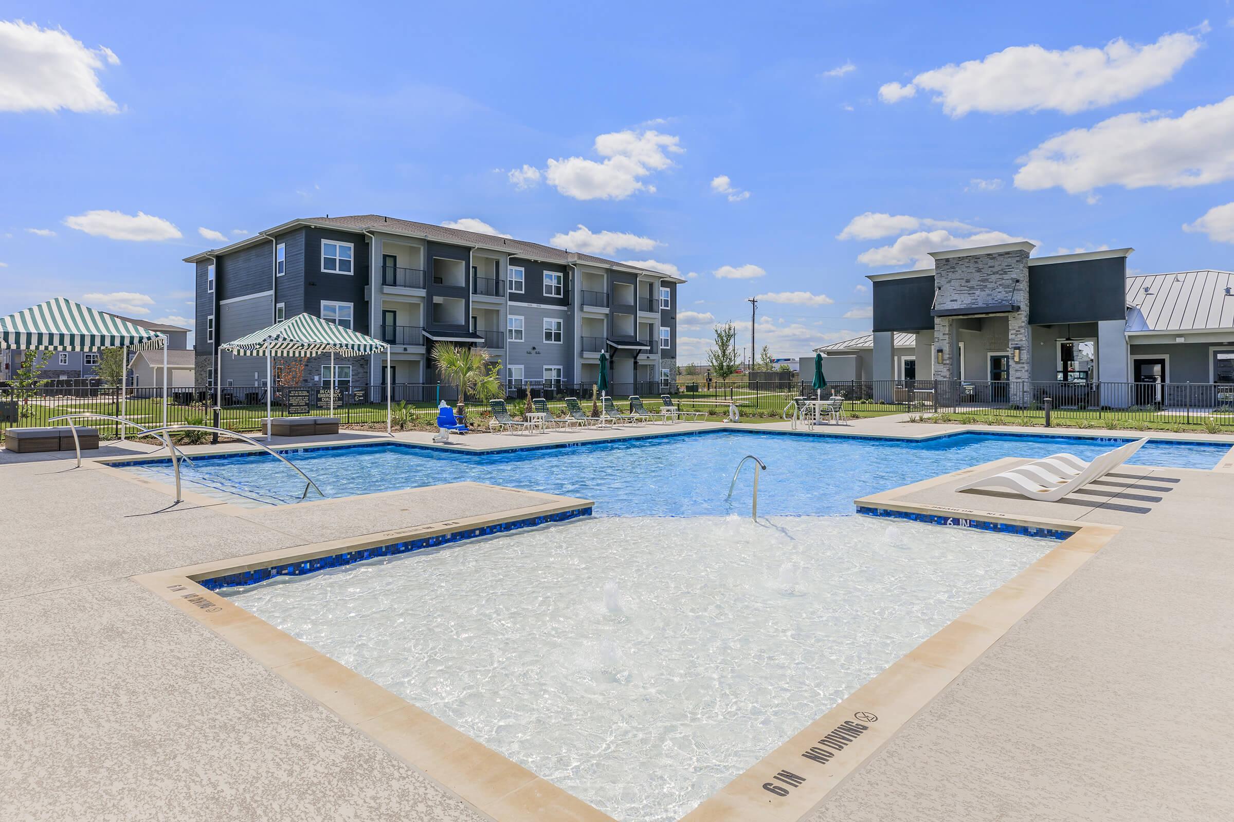 A bright outdoor pool area featuring two swimming pools, one with fountains, surrounded by lounge chairs and umbrellas. In the background, modern apartment buildings under a blue sky with fluffy white clouds provide a welcoming residential atmosphere.