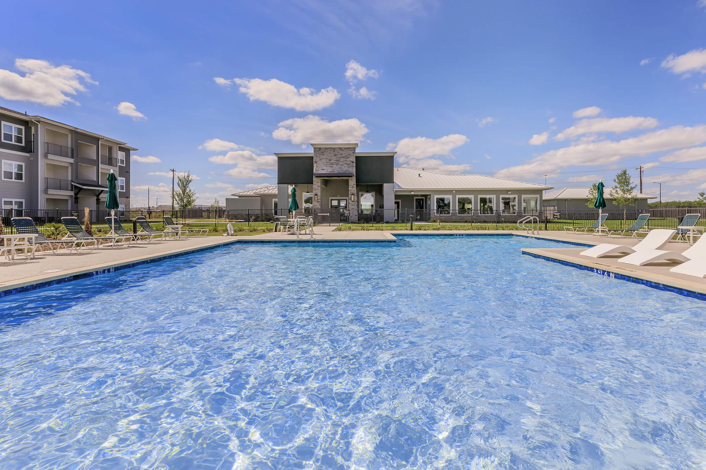 A bright blue swimming pool in the foreground, with lounge chairs arranged around it. In the background, a modern building features large windows and an inviting entrance, set against a clear blue sky with fluffy clouds. Lush greenery surrounds the area, creating a relaxing atmosphere.