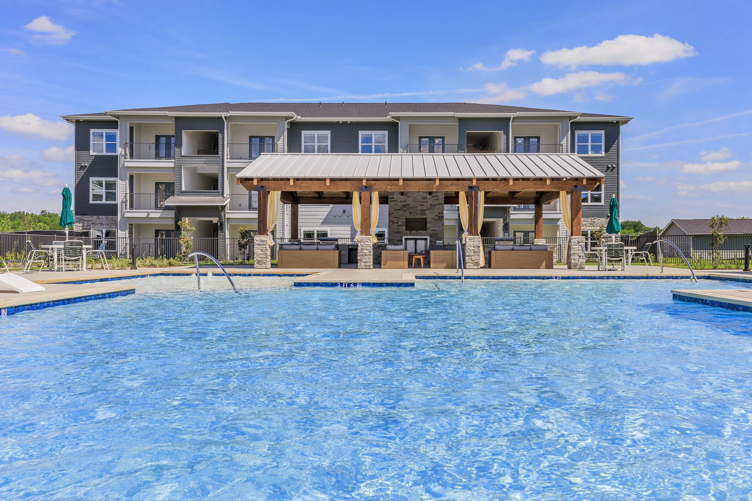 A spacious swimming pool in the foreground with clear blue water, surrounded by lounge chairs and umbrellas. In the background, a modern apartment building features balconies and multiple floors, with a shaded patio area providing seating. Bright blue sky and fluffy clouds complete the scene.