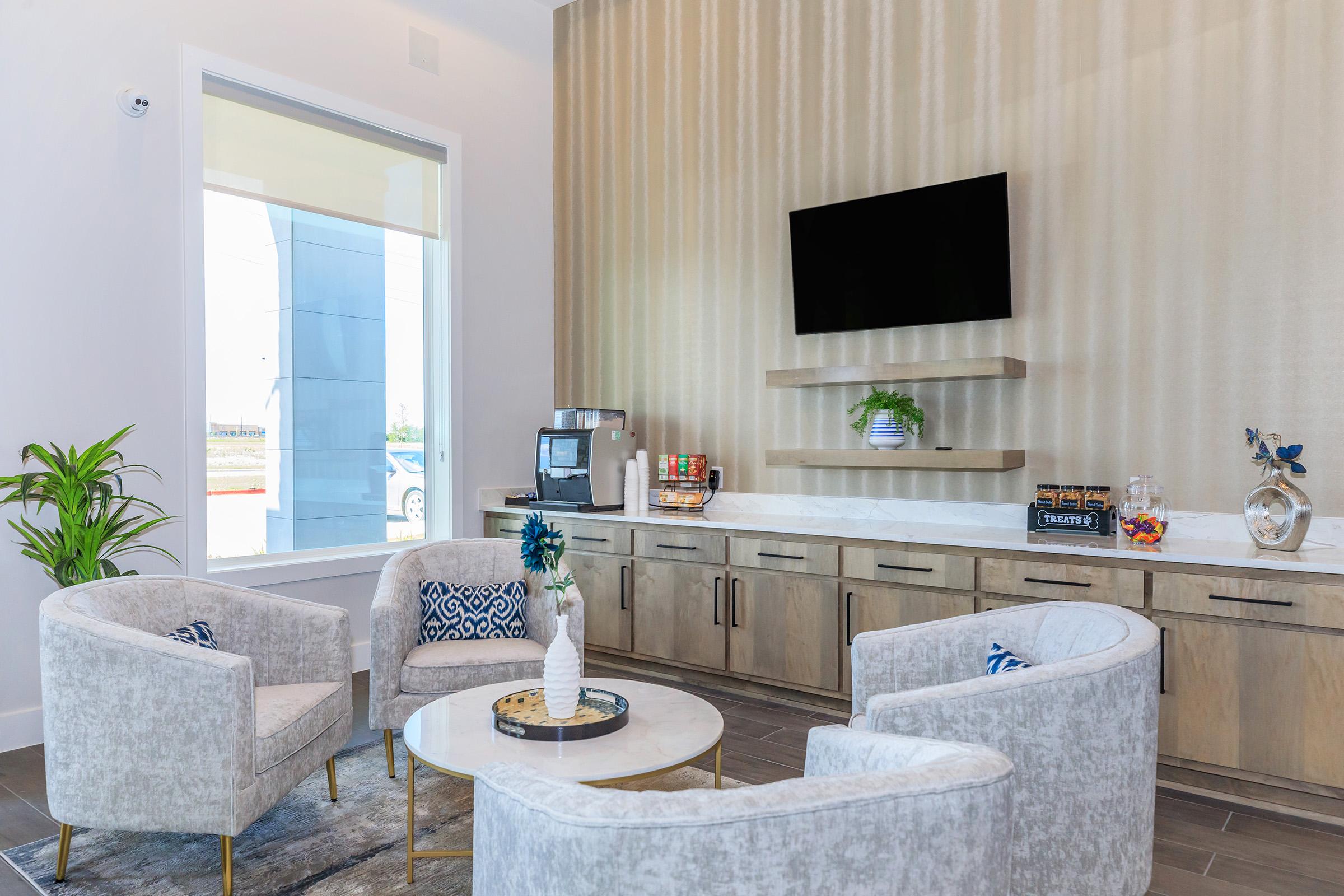 A modern lounge area featuring three light gray armchairs around a round table. There’s a small plant on the table, a coffee maker on a console below a wall-mounted TV, and decorative items on the shelves. Large windows let in natural light, enhancing the cozy atmosphere.