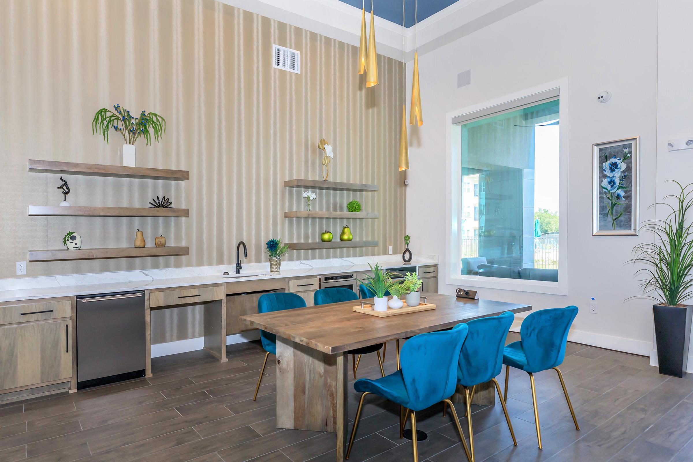 Modern kitchen and dining area featuring a sleek wooden table surrounded by blue chairs, wall shelves with decorative items, and large windows allowing natural light. Stylish pendant lights hang from the ceiling, enhancing the contemporary design.