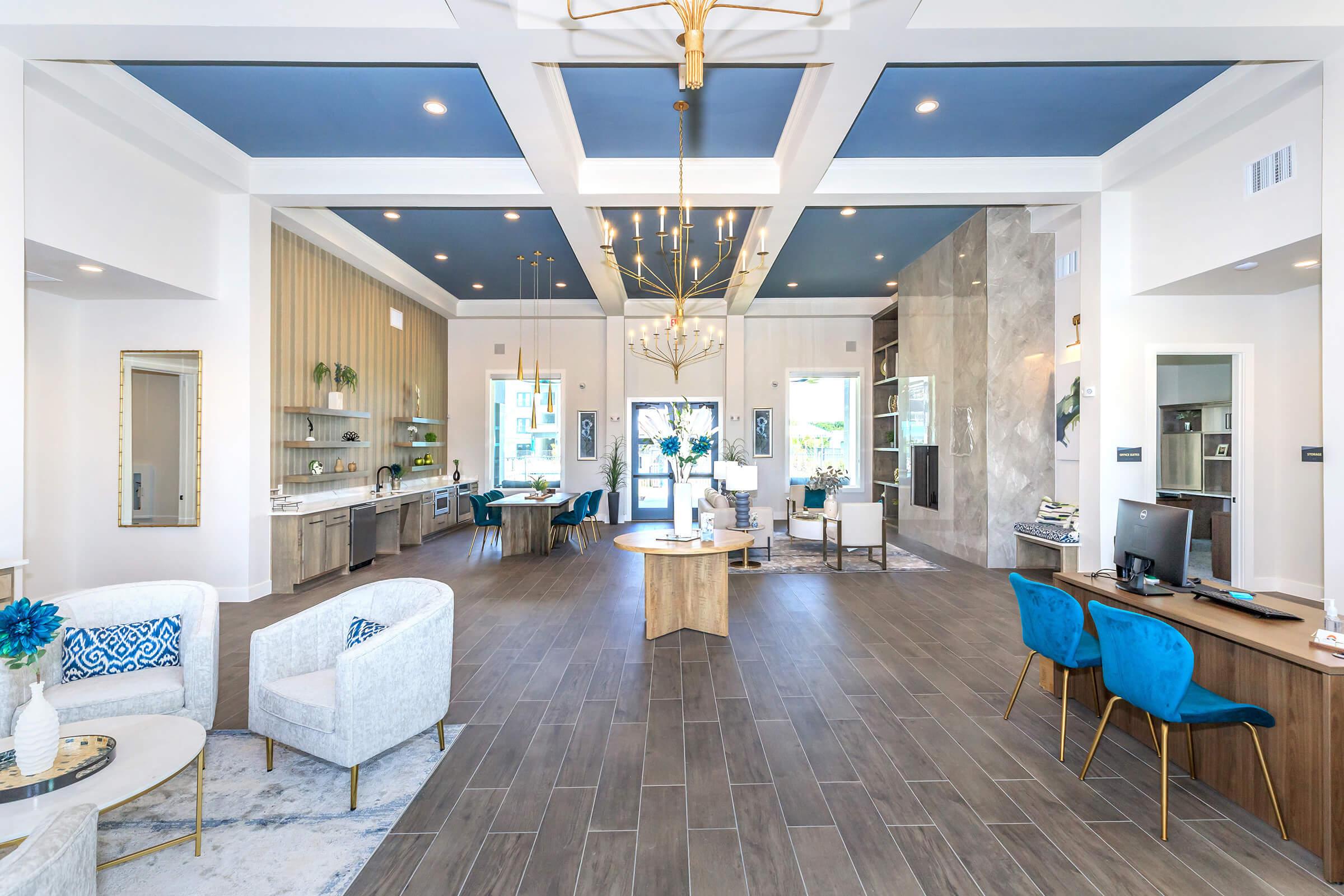 Modern lobby area featuring stylish furniture, including white chairs and blue accent chairs. There are decorative shelves and a large chandelier, all set against a backdrop of wood and marble finishes. Natural light enters through large windows.
