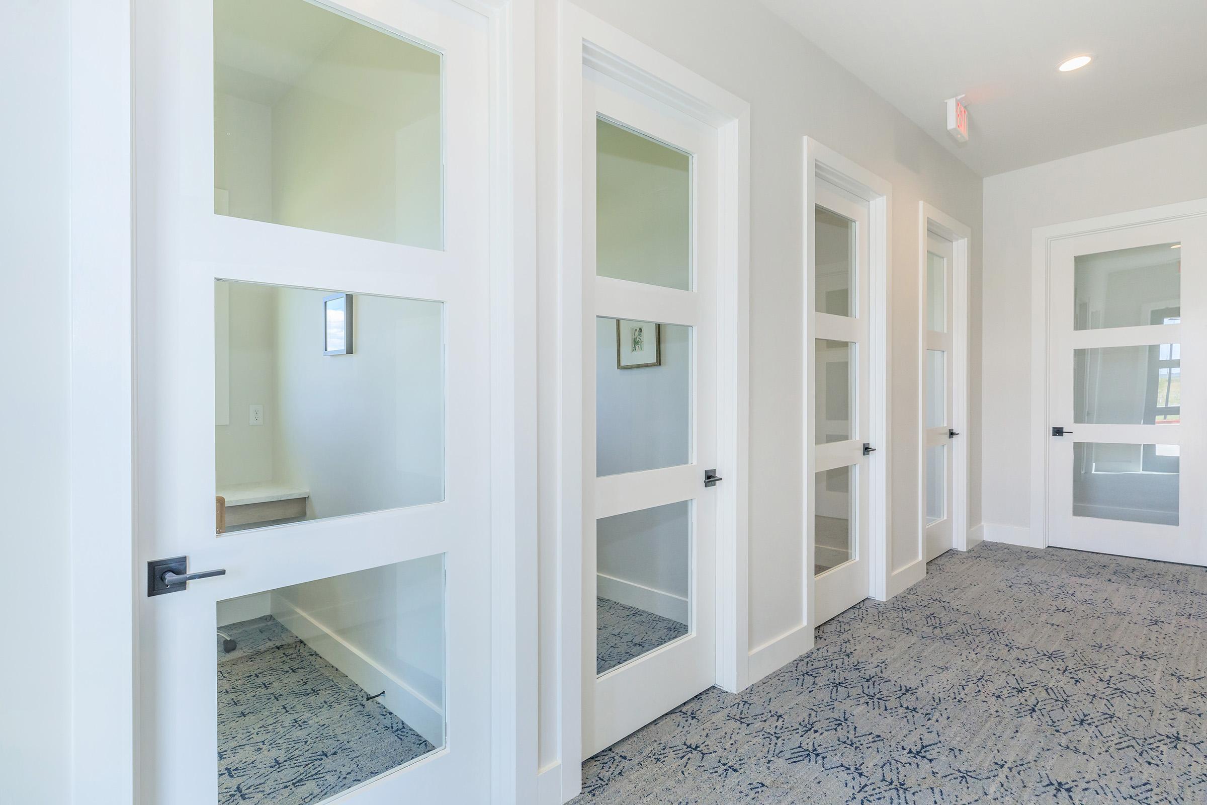 A hallway featuring multiple frosted glass-panel doors leading to small rooms, with a light gray wall and patterned carpet. The space is bright and modern, showcasing a clean, minimalist design.