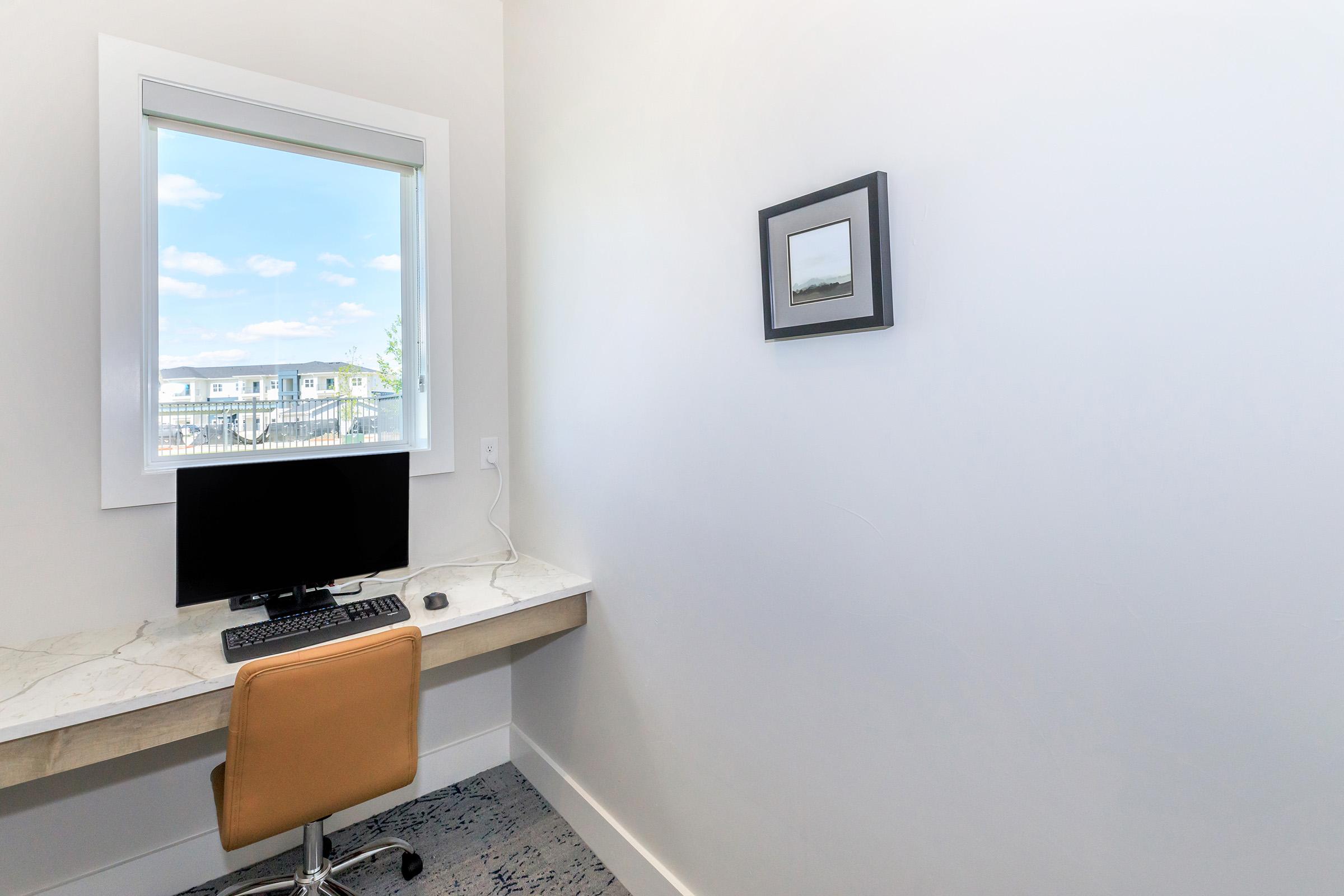 A tidy workspace featuring a computer monitor, keyboard, and mouse on a marble desk. An orange office chair is positioned in front of the desk. A window offers a view of the outside with blue sky and clouds. A framed picture is mounted on the wall opposite the desk, adding a touch of decor to the minimalist room.