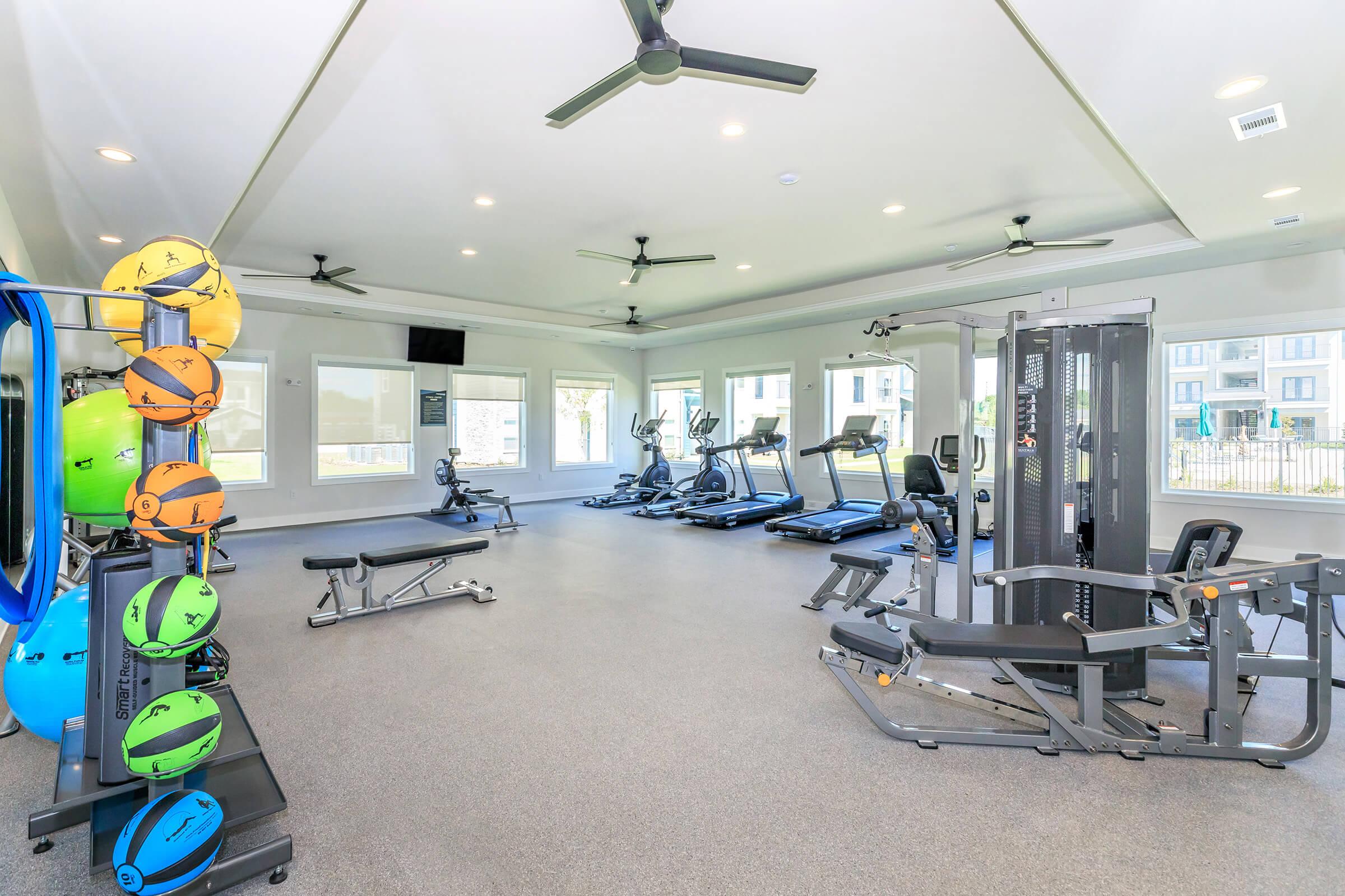 A modern gym interior featuring various exercise equipment, including treadmills, elliptical machines, a weight machine, and free weights. The room is well-lit with windows and has ceiling fans, creating an open and inviting space for fitness activities.