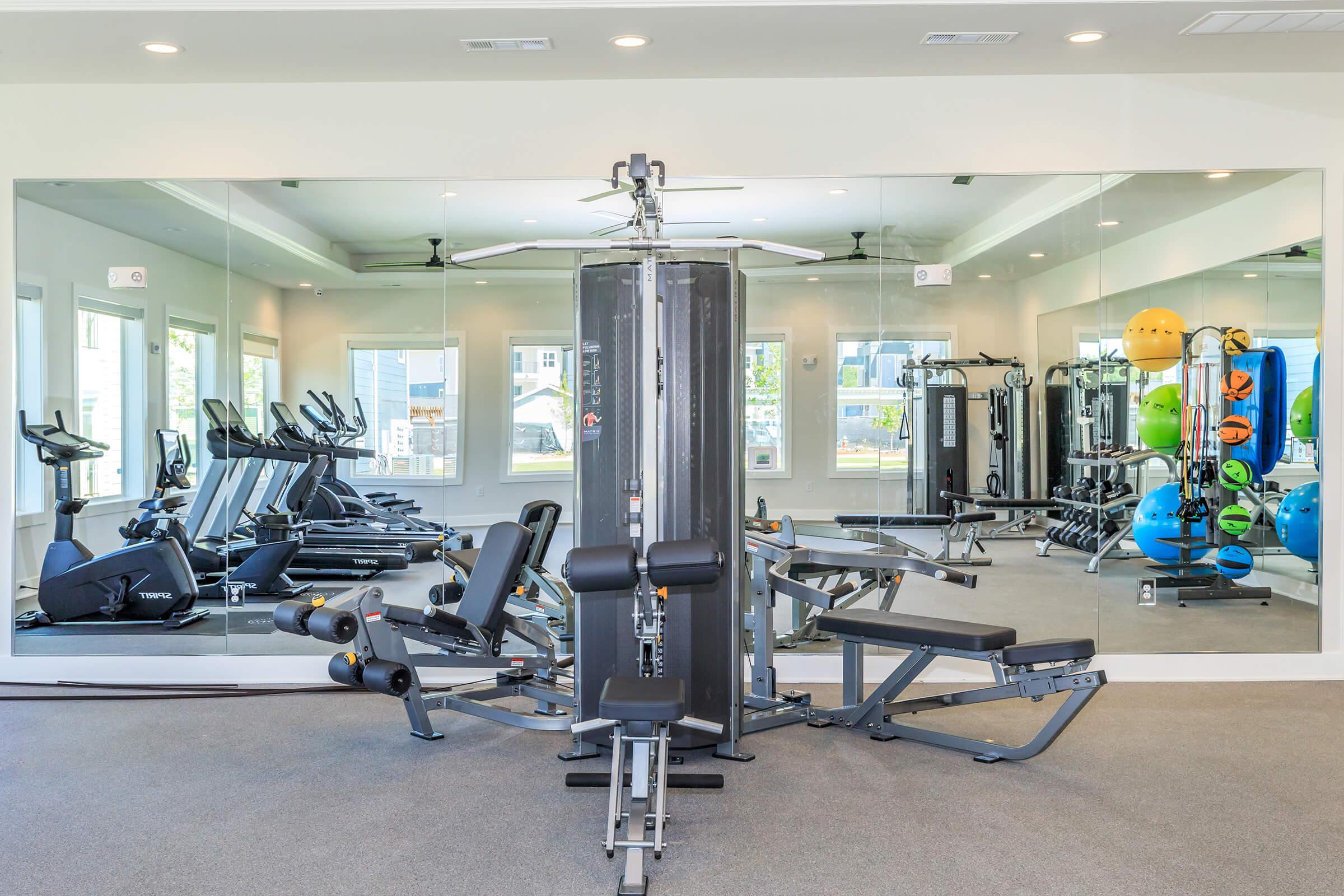 A well-lit gym featuring exercise equipment including a multi-gym machine, exercise bikes, and weights. Large mirrors line the walls, reflecting the equipment and creating an open, spacious feel. The room is modern and inviting, with natural light streaming in through windows.
