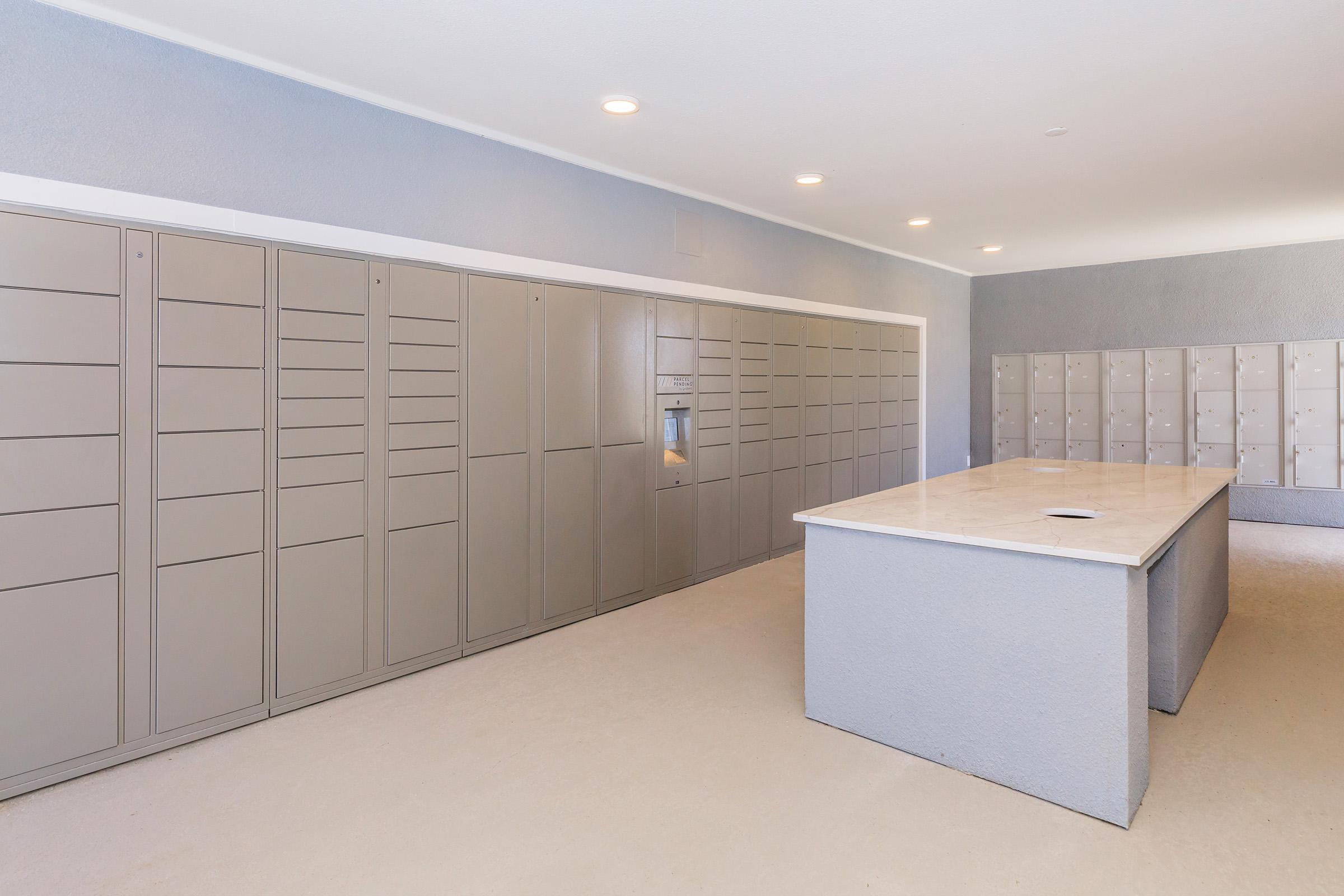 A modern mailroom featuring rows of gray lockers against a light-colored wall. There is a central reception table made of stone, providing a clean and organized space for collecting and distributing mail. The room is well-lit with recessed lighting, creating a bright and open atmosphere.