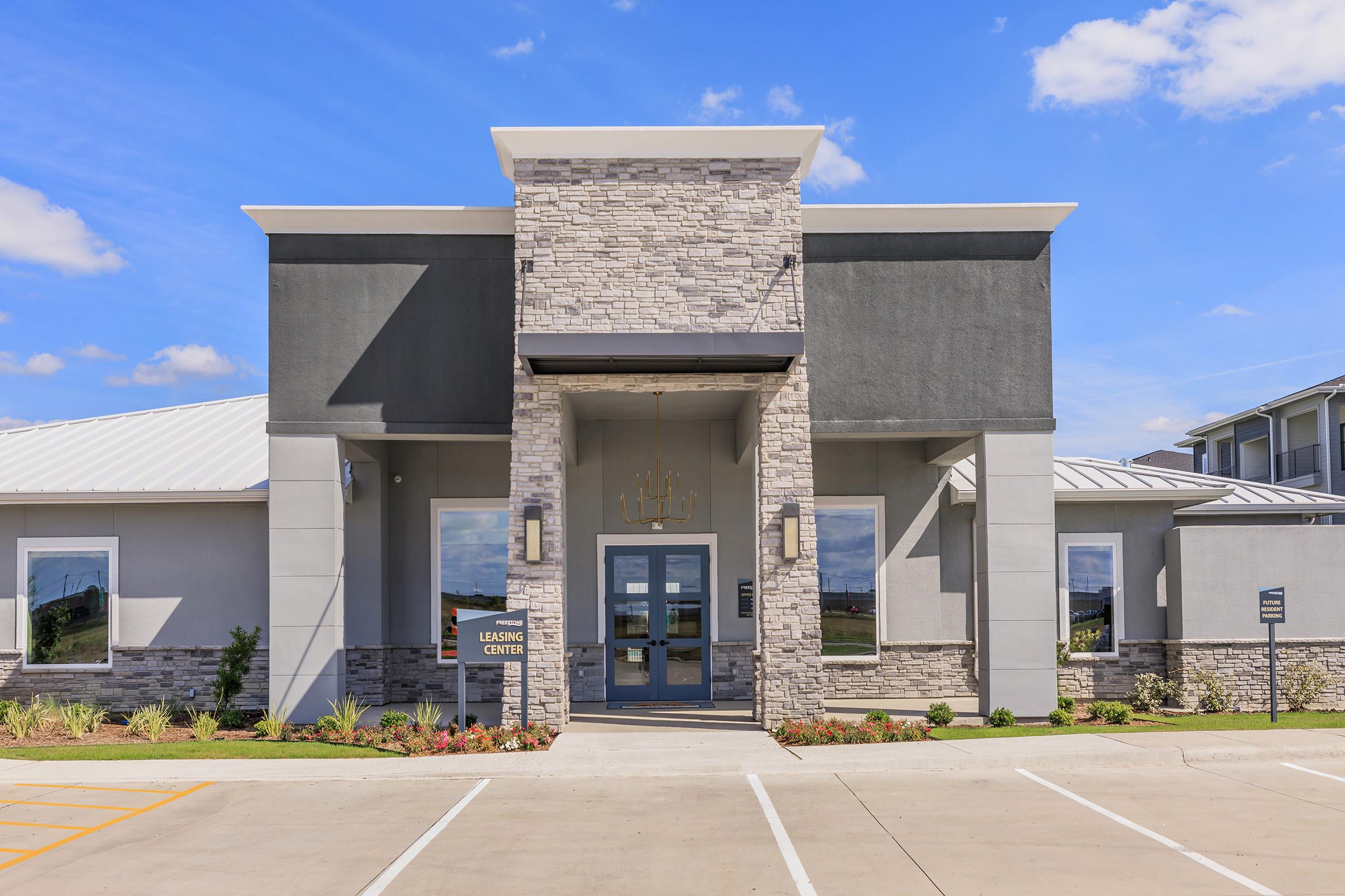 A modern leasing center with a stone and gray exterior, large glass front doors, and a light fixture visible through the entrance. The building is surrounded by well-maintained landscaping under a clear blue sky. Parking spaces are marked in front of the entrance, leading to an inviting atmosphere.
