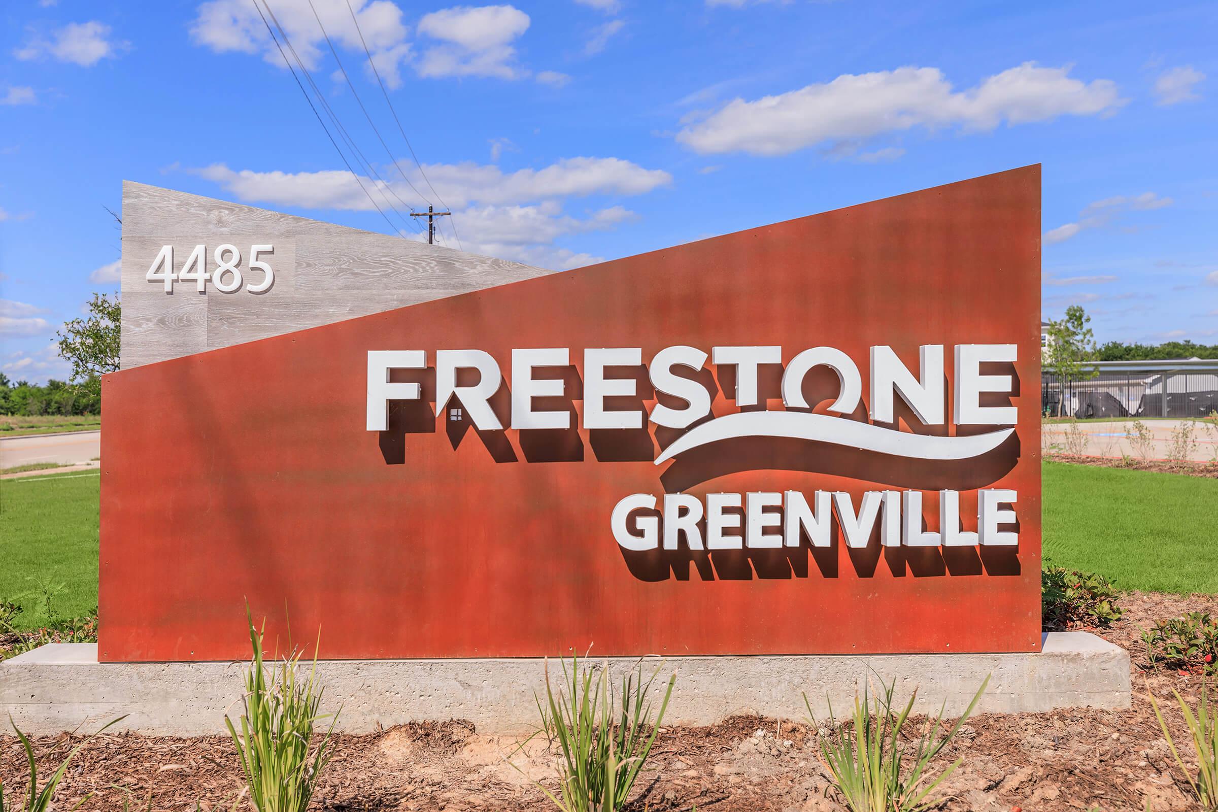Sign displaying "FREESTONE" in large white letters, with "GREENVILLE" underneath in smaller text. The sign is mounted on a modern, rust-colored background, with a clean design. It is situated in an outdoor area with green grass and a blue sky. The location number "4485" is also visible.