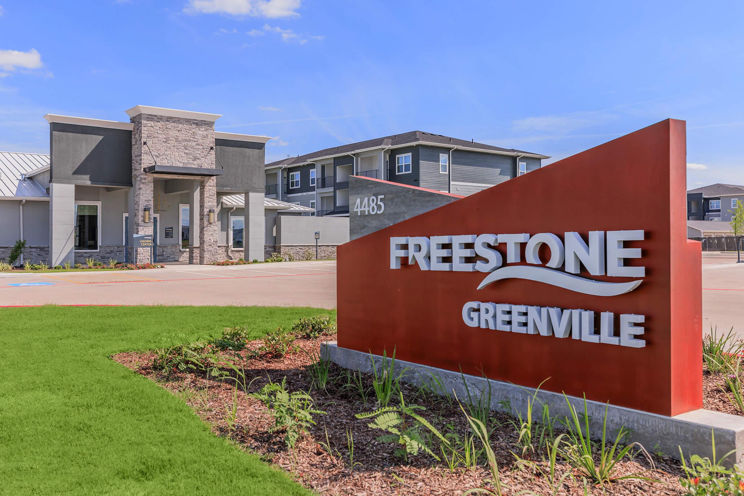 Exterior view of the Freestone Greenville apartment complex, featuring a modern entrance with a stone façade and a prominent red sign displaying "FREESTONE" and "GREENVILLE." Landscaping includes green grass and plants in the foreground with a clear blue sky in the background.