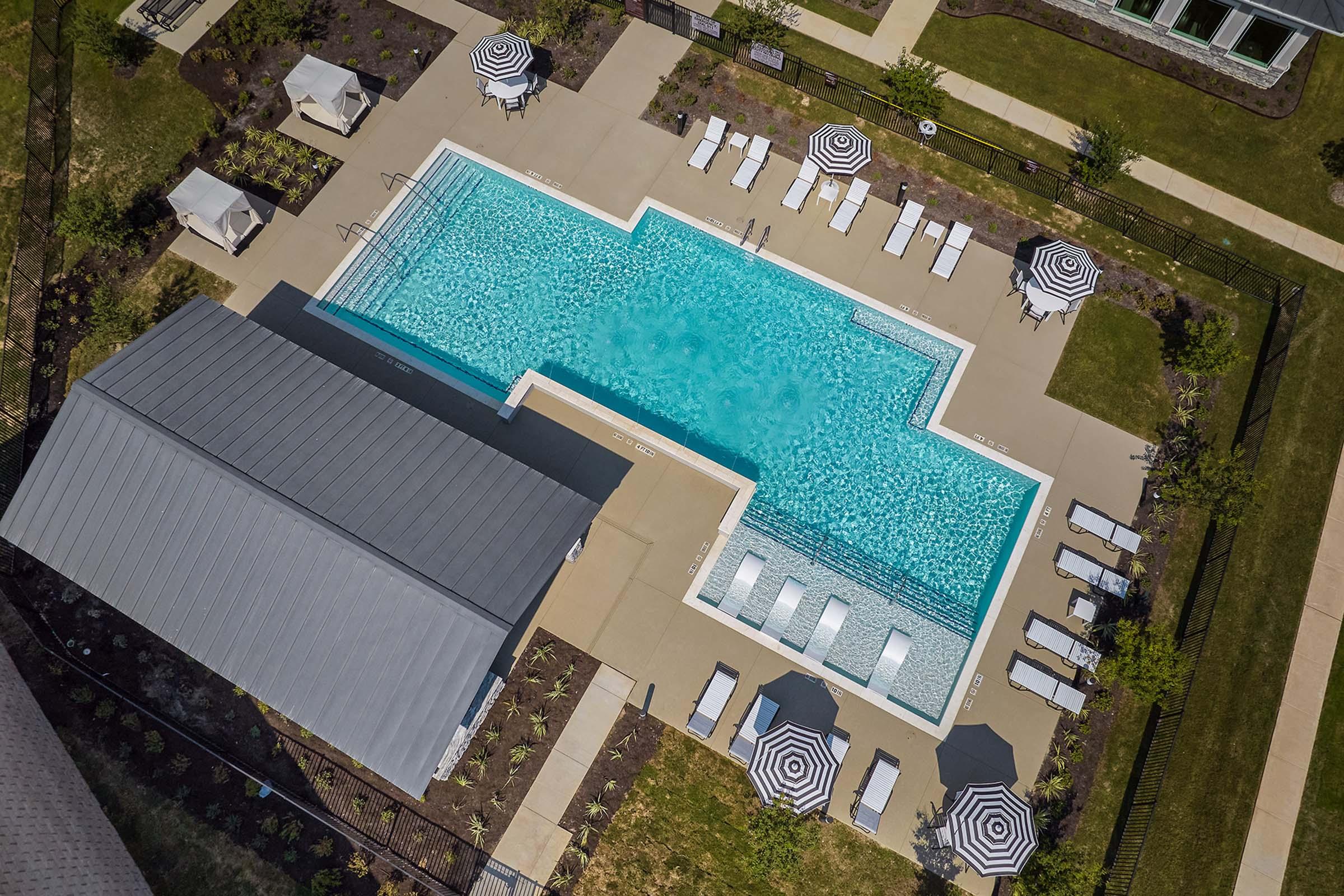 Aerial view of a modern swimming pool surrounded by lounge chairs and umbrellas. The pool has a unique shape, with clear blue water reflecting sunlight. Nearby, there are shaded seating areas under canopies, and well-maintained green grass adds to the outdoor relaxation space.