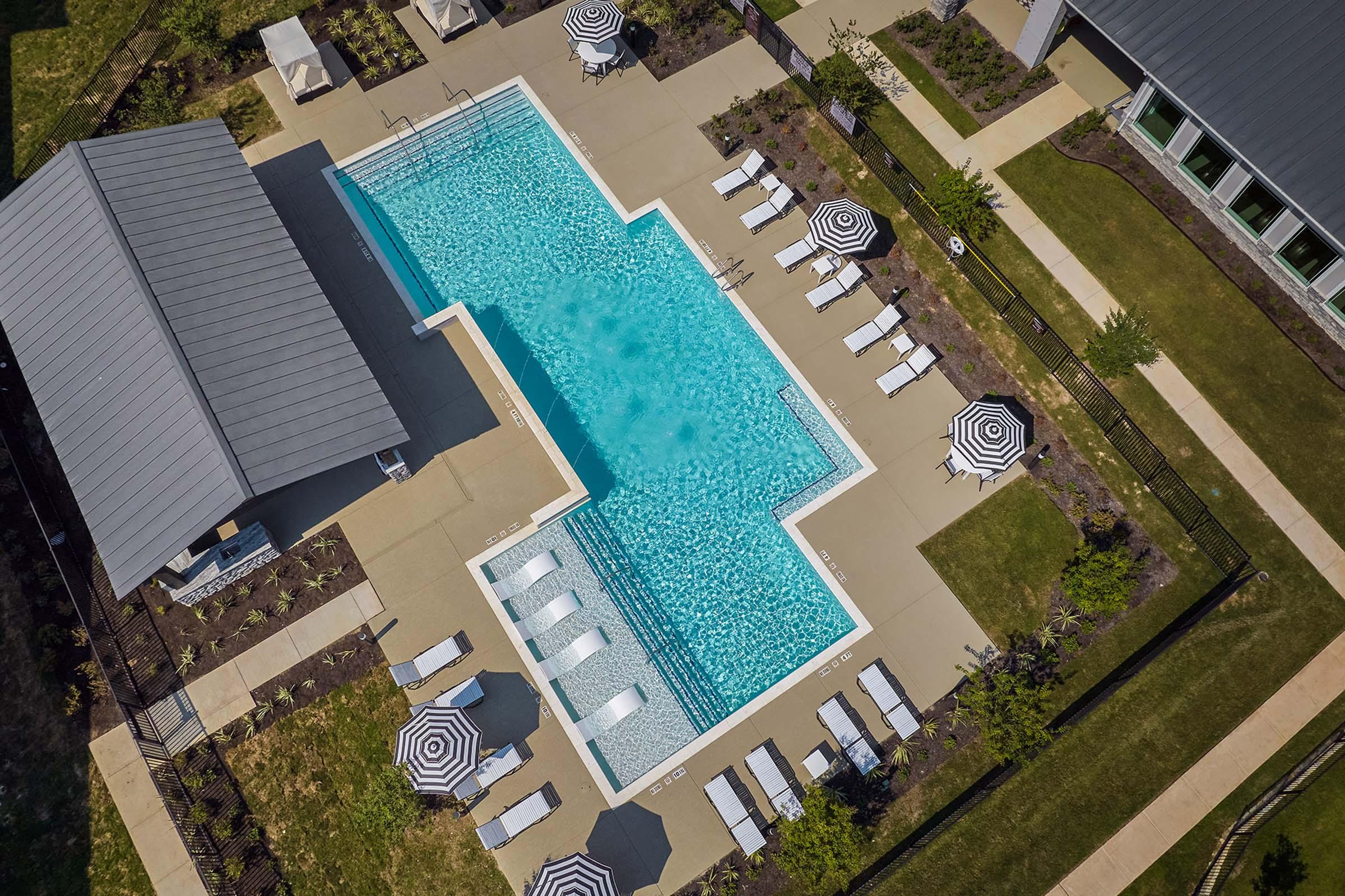 Aerial view of a swimming pool surrounded by lounge chairs and umbrellas. The pool features a clear blue water surface and is bordered by a concrete patio. Nearby, there are landscaped areas and a building with a gray roof, creating a relaxing outdoor environment.