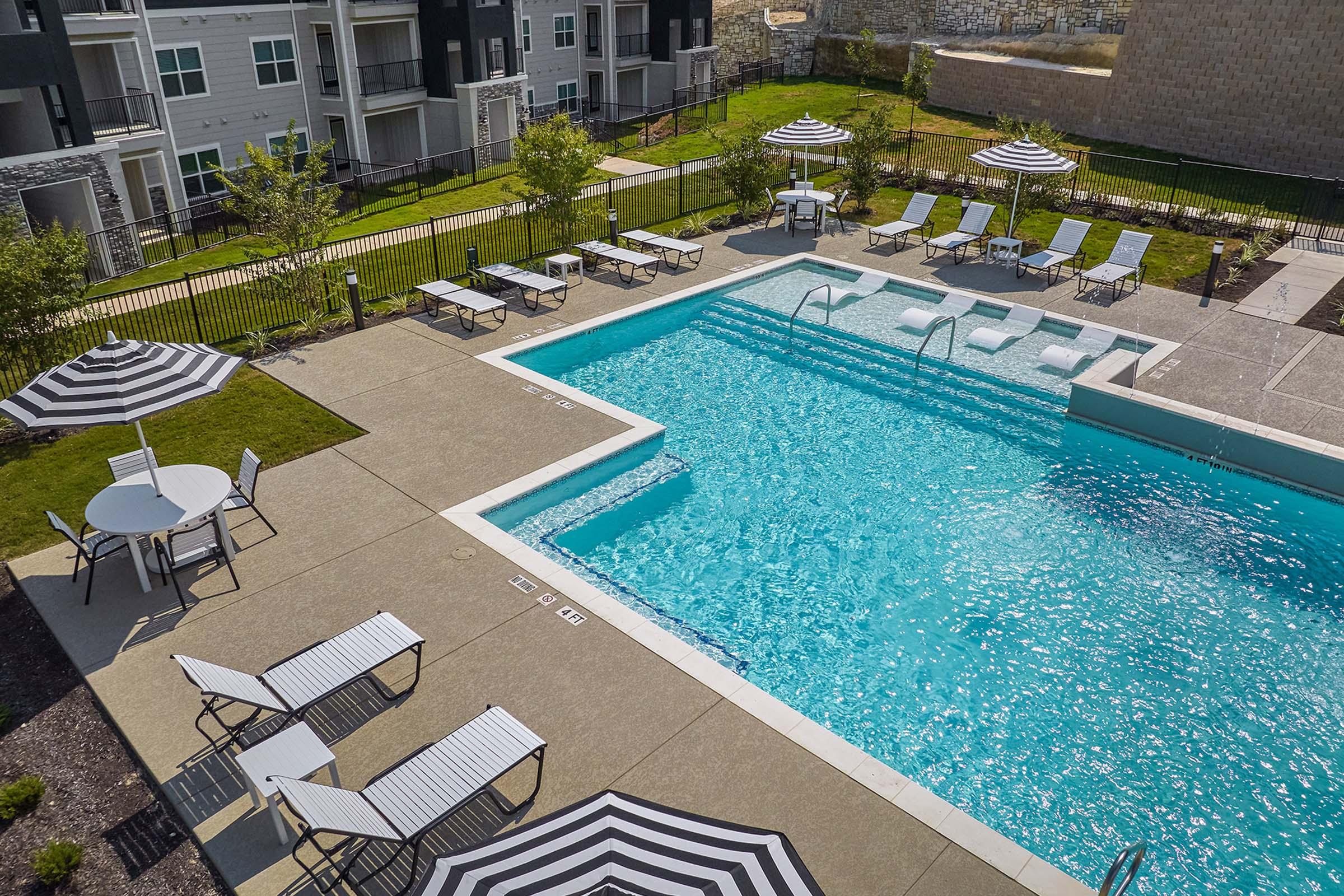 A spacious outdoor swimming pool surrounded by lounge chairs and tables, featuring striped umbrellas for shade. The pool area is situated within a well-maintained residential complex, with green grass and modern buildings visible in the background.