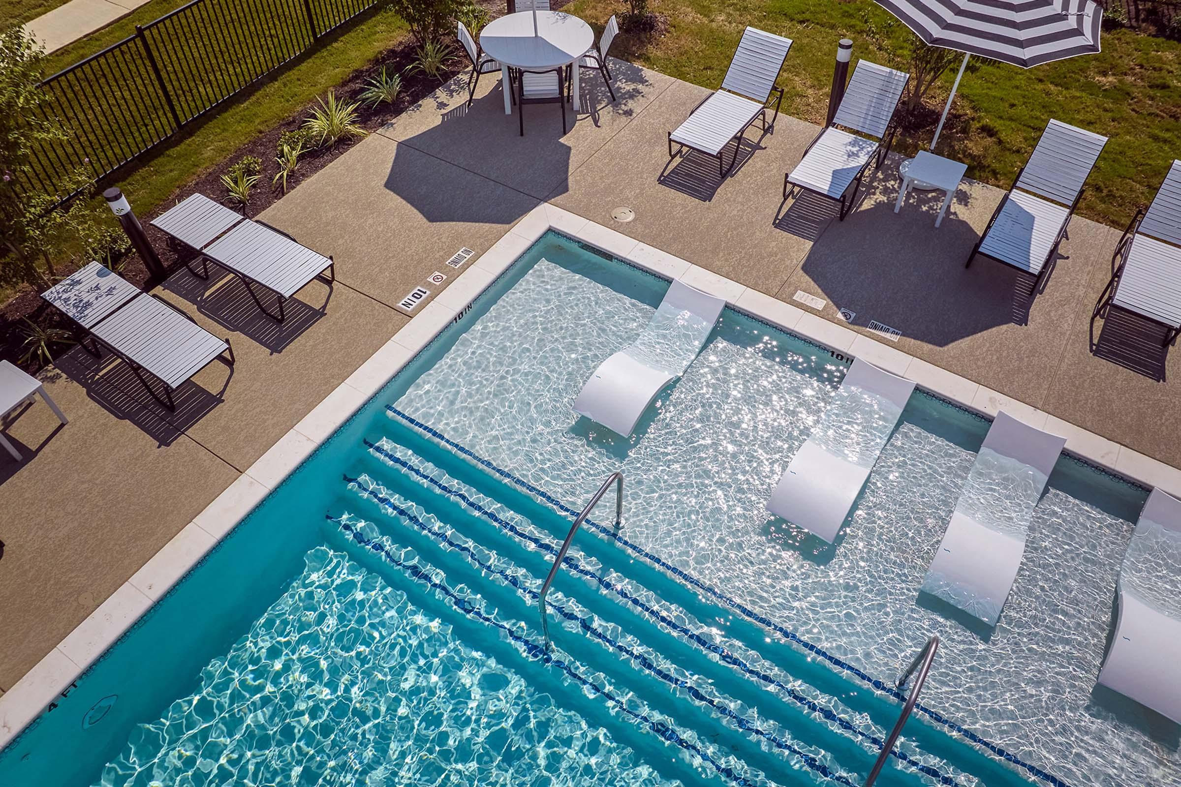 Aerial view of a swimming pool with lounge chairs and umbrellas surrounding it. The pool features in-water loungers for relaxation, and neatly arranged tables and chairs on a patio area. Lush greenery and a fence are visible in the background, creating a serene outdoor setting.