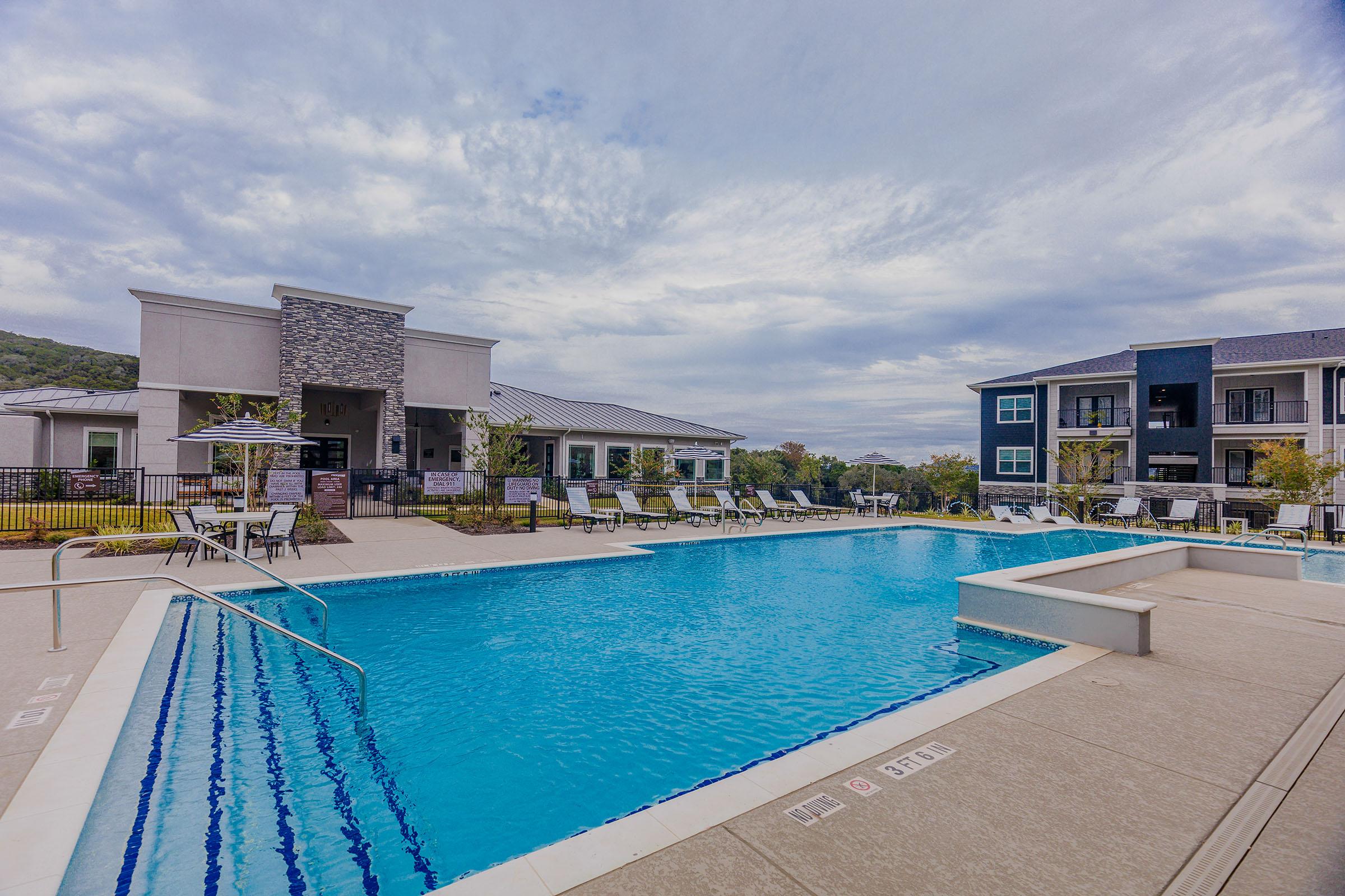 A modern apartment complex featuring a large swimming pool surrounded by lounge chairs. There are umbrellas and tables for relaxation, with well-maintained landscaping in the background. The sky is slightly cloudy, creating a serene atmosphere for residents.