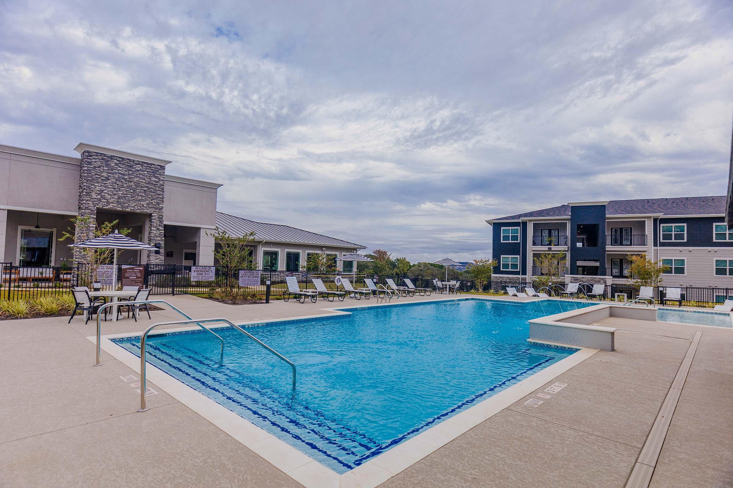 A swimming pool in an outdoor area surrounded by lounge chairs and tables. In the background, there are modern apartment buildings under a cloudy sky. The setting conveys a relaxing atmosphere with greenery around the pool area.