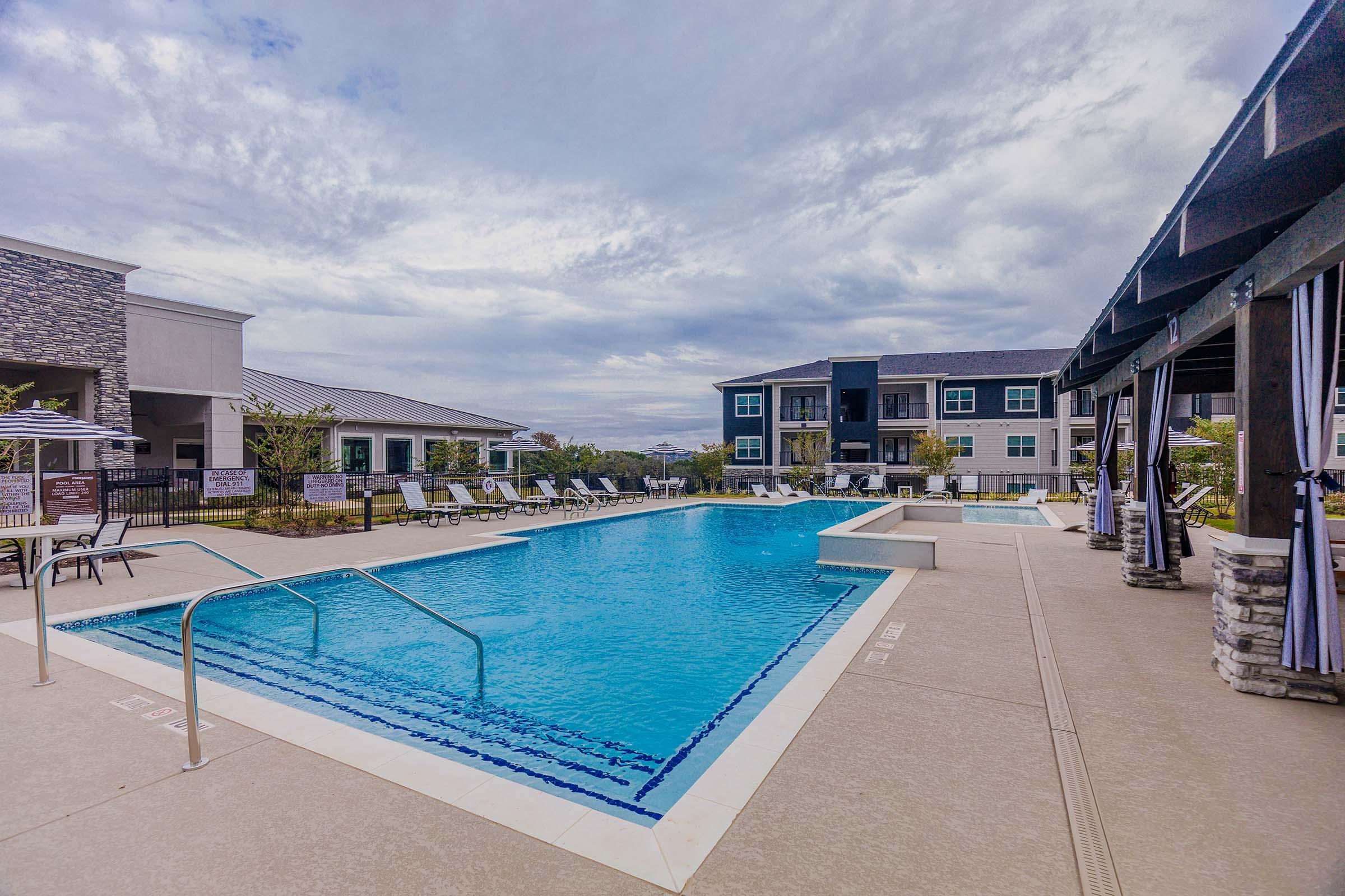 A serene outdoor swimming pool surrounded by lounge chairs, with a stylish building in the background. The pool area features a modern design, umbrellas for shade, and lush greenery nearby, under a partly cloudy sky. Ideal for relaxation and leisure activities.