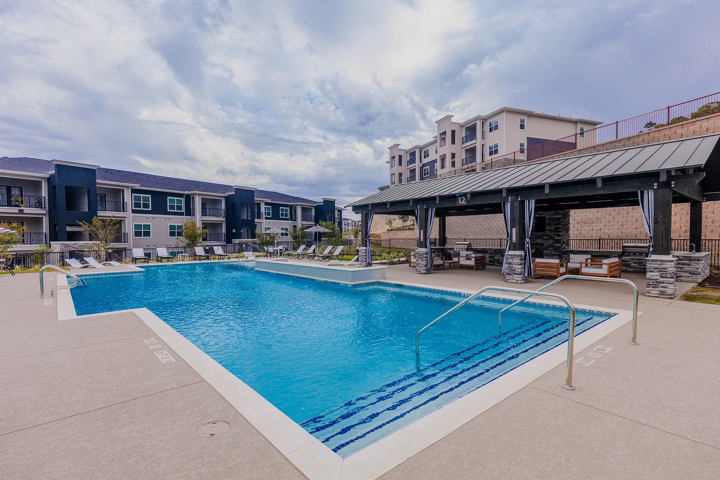 A clear blue swimming pool surrounded by lounge chairs, with a shaded sitting area featuring wooden furniture. In the background, multiple modern apartment buildings are visible under a cloudy sky. The setting is inviting and designed for relaxation.