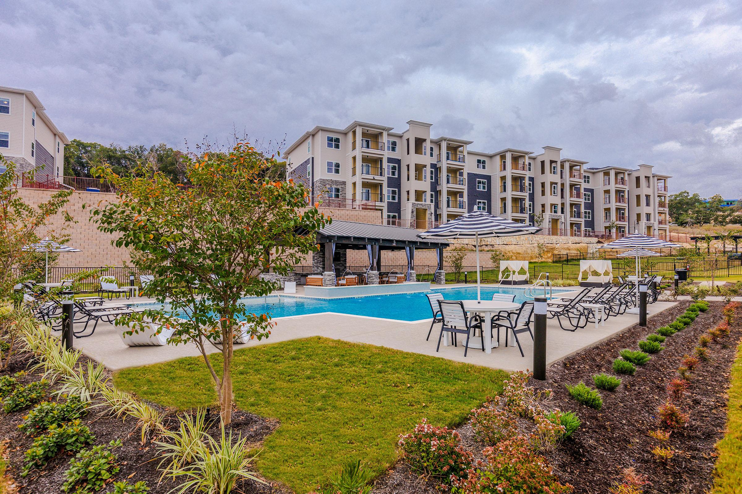 A modern residential pool area featuring a large swimming pool surrounded by lounge chairs. The landscape includes neatly trimmed shrubs and flower beds. In the background, there are several multi-story apartment buildings, with a cloudy sky above creating a serene atmosphere.