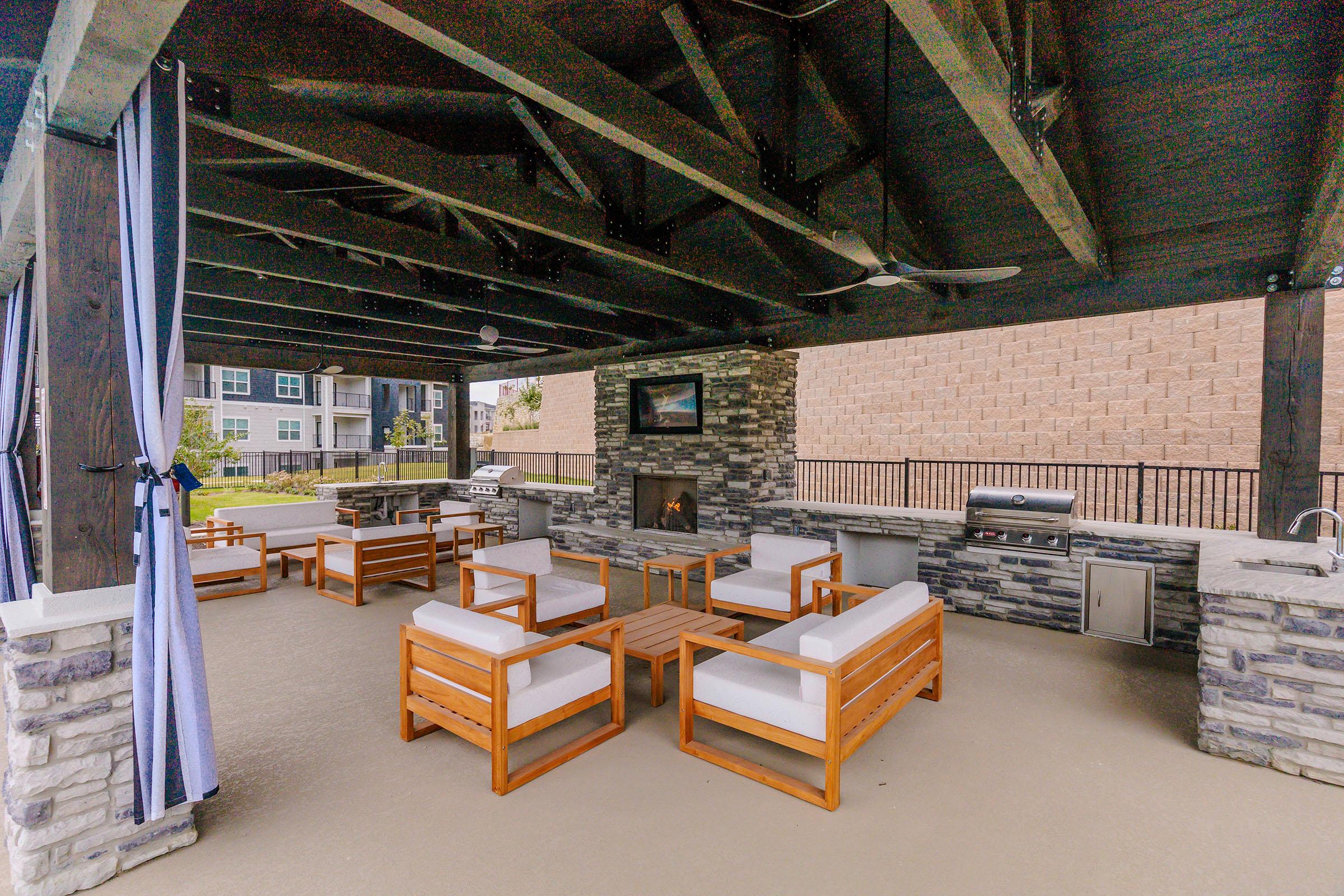 Outdoor lounge area under a canopy featuring comfortable wooden seating with white cushions, a central coffee table, an outdoor fireplace, a mounted TV, and a grill setup. The space has stone accents and is set against a backdrop of modern buildings.