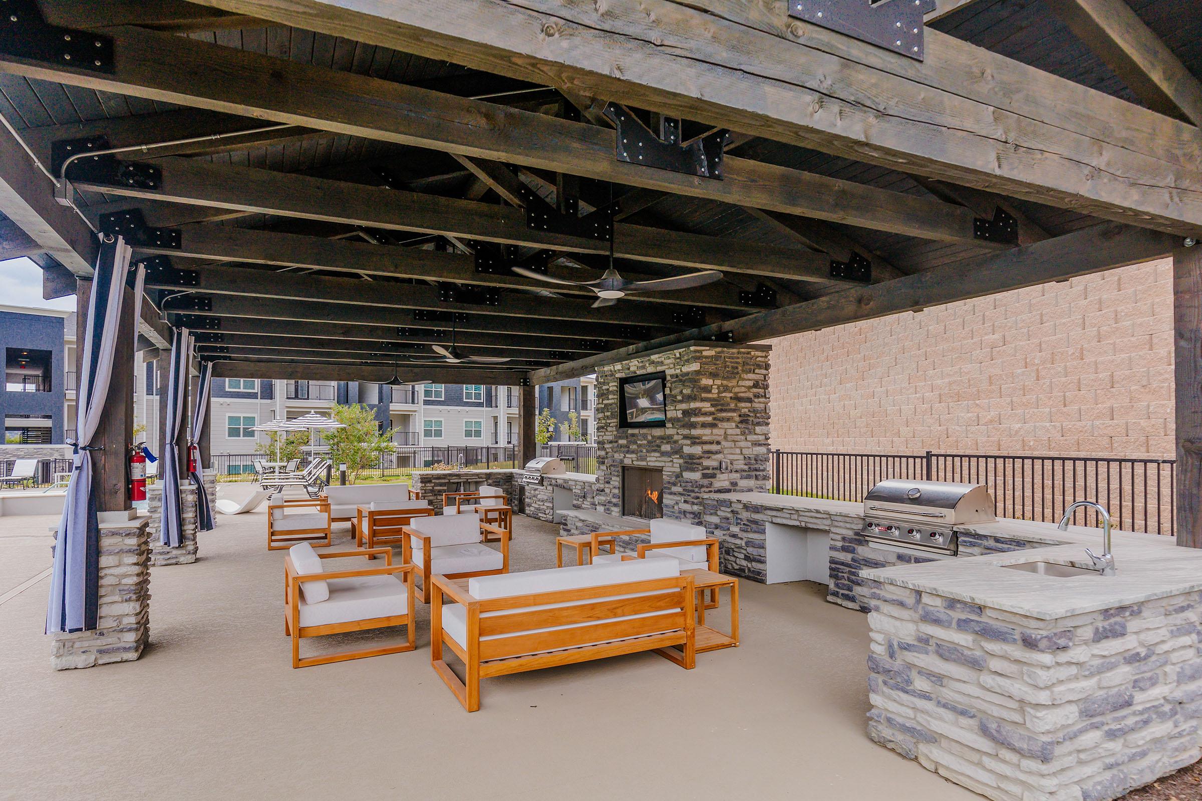 A spacious outdoor lounge area featuring wooden seating arrangements and stone accents. There’s a built-in grill and countertop, a stone fireplace, and a ceiling fan, all under a large wooden canopy. In the background, there are modern apartment buildings and landscaped greenery.