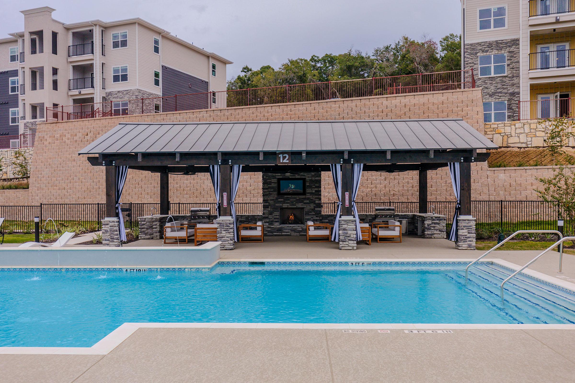 A serene outdoor pool area featuring a spacious swimming pool with a swim-up ledge. In the background, there’s a covered seating area with modern lounge furniture, surrounded by neatly landscaped greenery and an apartment building. The overcast sky adds a calm atmosphere to the setting.