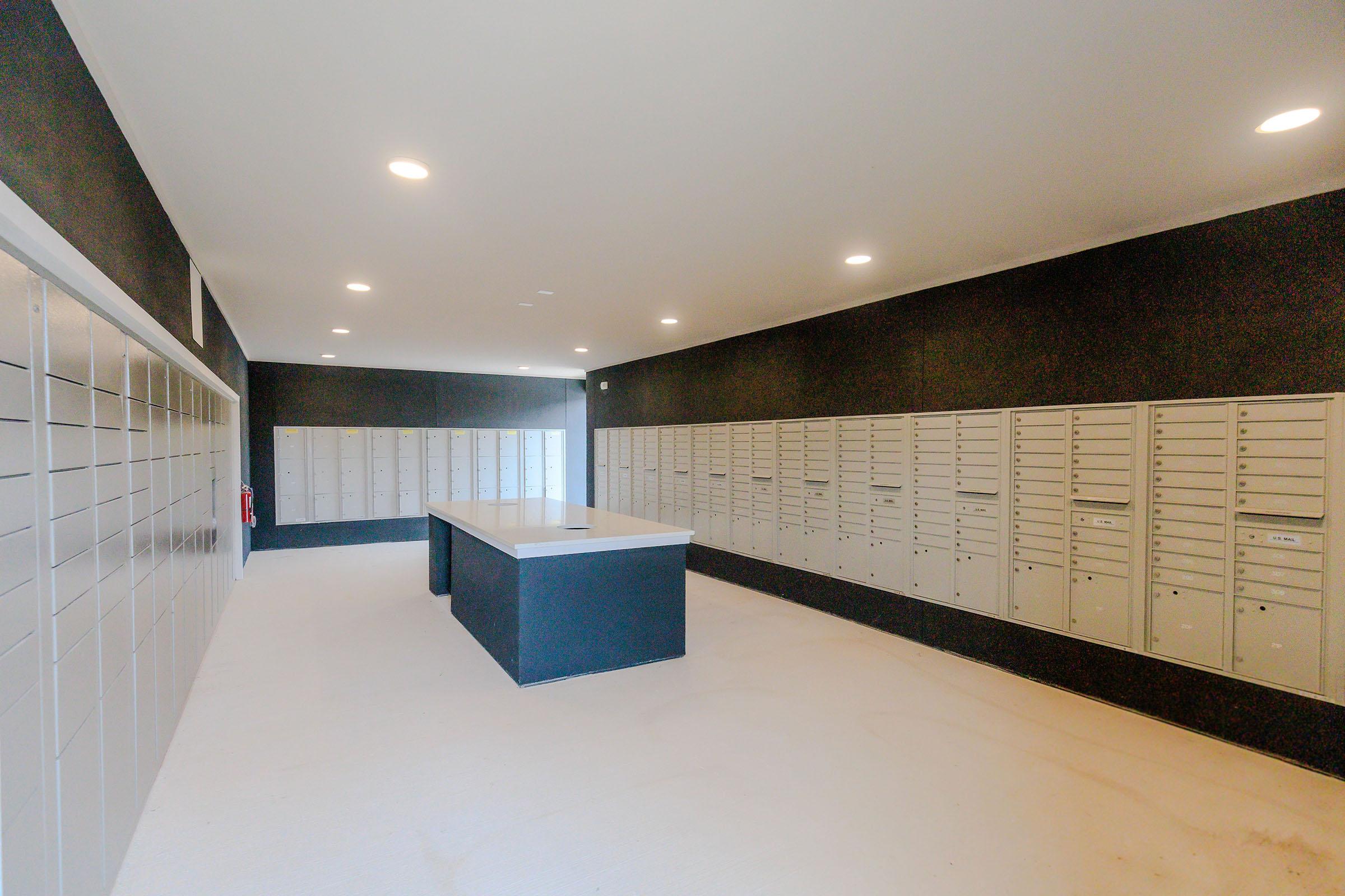 Interior of a modern mailroom featuring gray mailboxes arranged on two walls, with a central table. The walls are painted black, and overhead lights provide bright illumination. The space appears clean and organized, designed for receiving and distributing mail securely.