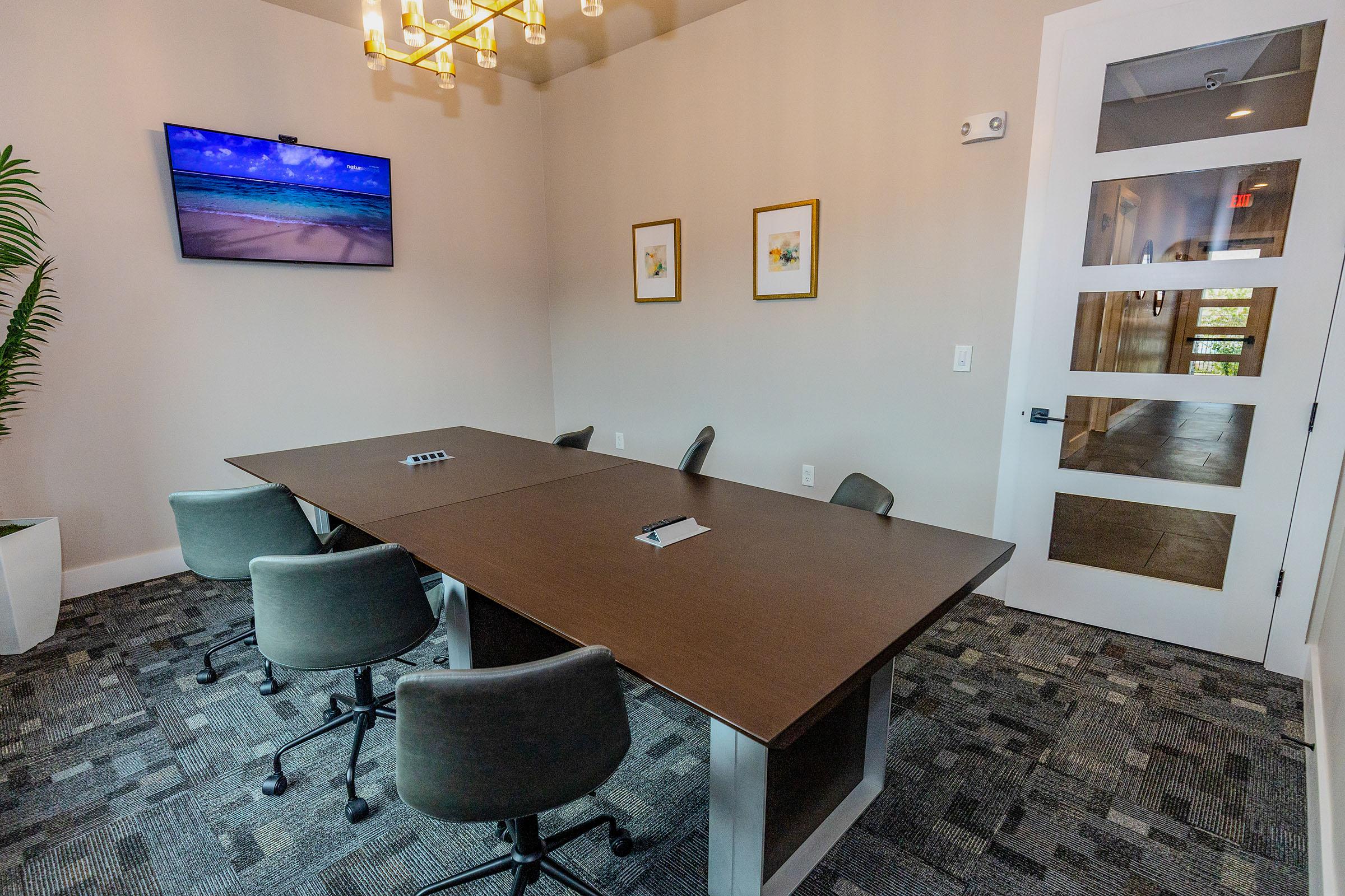 A modern conference room featuring a large wooden table surrounded by six ergonomic chairs. A flat-screen TV mounted on the wall displays a scenic beach image. The room has neutral-colored walls, framed artwork, and a plant, creating a professional, inviting atmosphere.