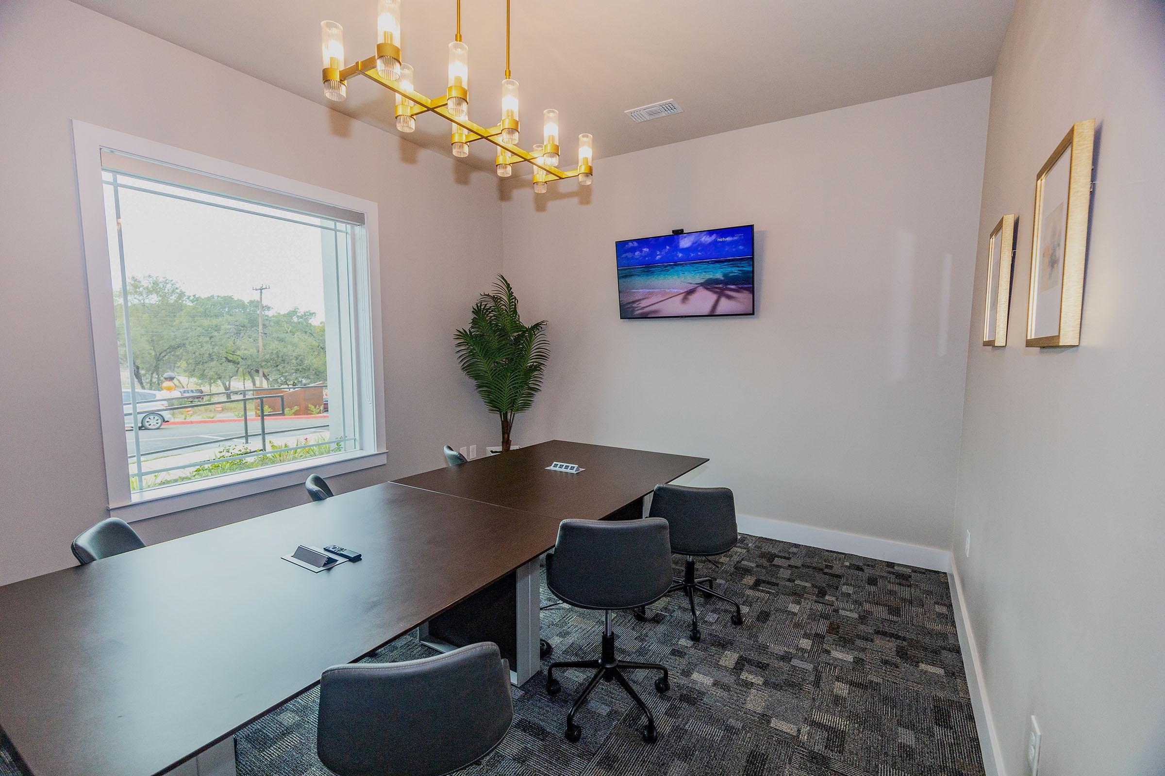 A modern conference room featuring a rectangular table surrounded by black chairs, a large window with natural light, a television displaying a beach scene, decorative framed artwork on the walls, and a potted plant, all set in a light-colored space with a contemporary chandelier.