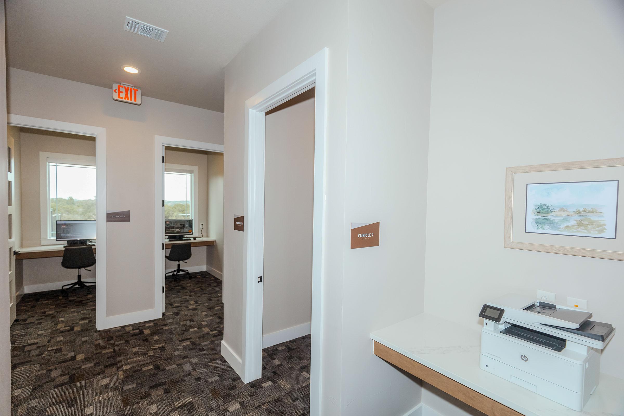 A well-lit office space featuring a corridor with two office rooms on the left and a printer on a desk on the right. Each office has a window and a desk with a chair. The walls are painted in neutral tones, and there is a piece of artwork hanging near the printer. A ceiling light and an exit sign are visible above.