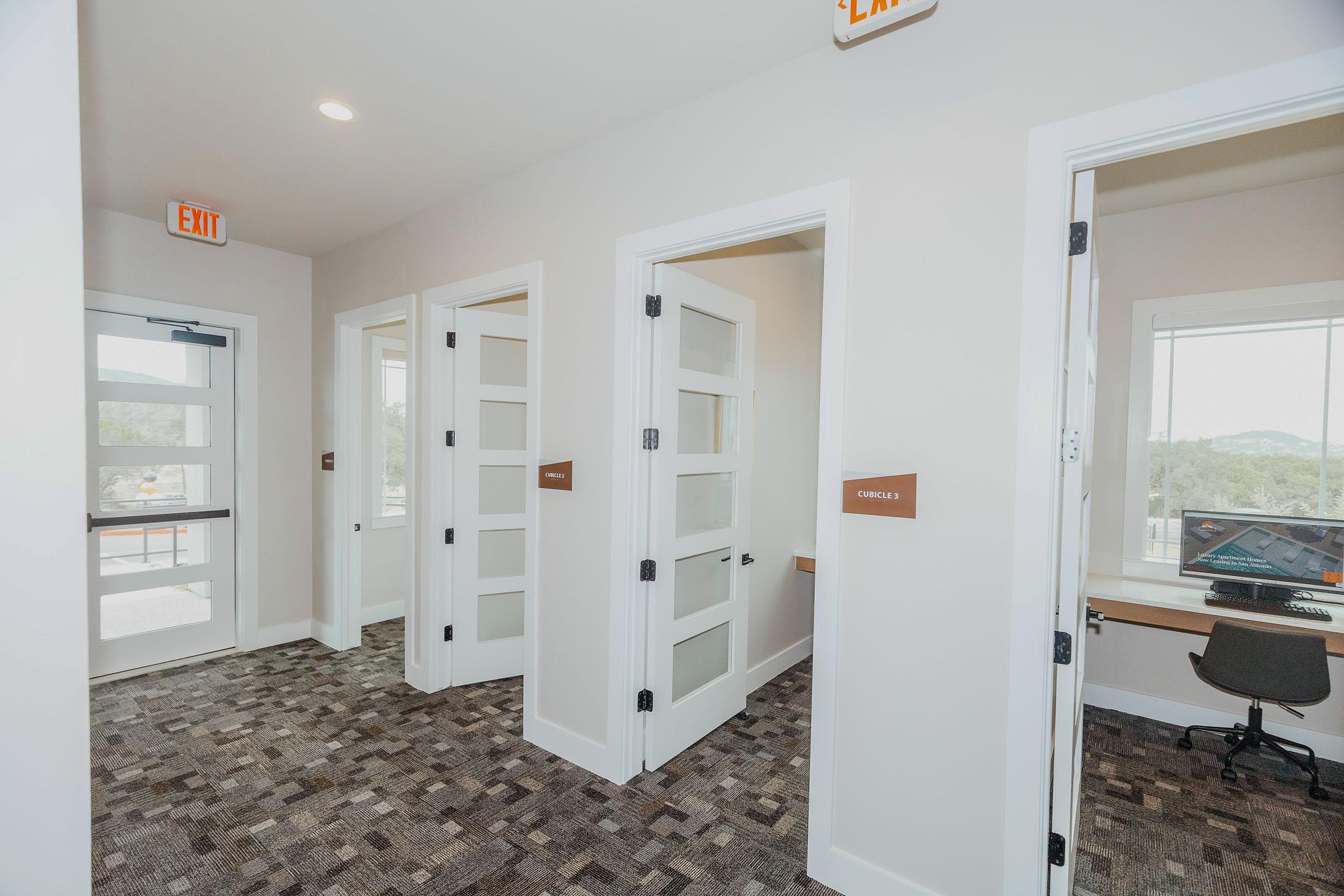 A well-lit hallway in a modern building, featuring several doors with frosted glass panels. On the left, there is an exit door with an emergency sign above. The adjacent doors are labeled and lead to small offices or rooms, with a workspace visible through a window at the end of the corridor. The floor has a patterned carpet.