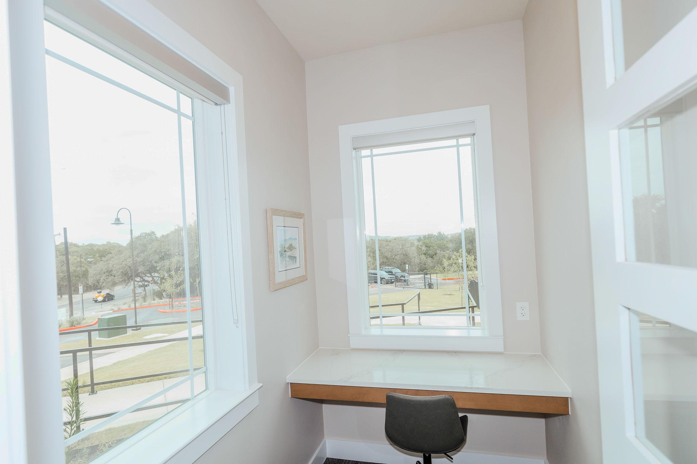 Bright interior corner with large windows allowing natural light, featuring a small desk with a chair. Decor includes a framed picture on the wall. Outside, trees and a road are visible through the windows. The design is modern and minimalistic, creating a calm workspace environment.