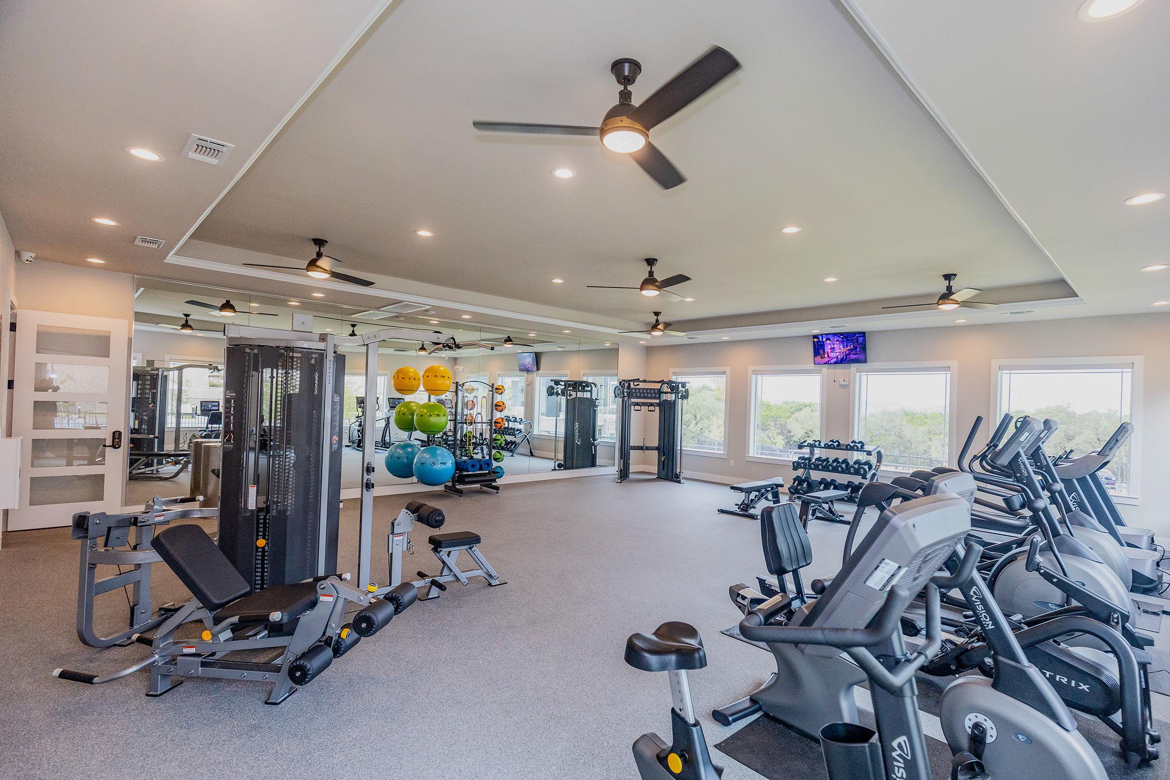 A spacious gym featuring various exercise equipment, including cardio machines, weights, and resistance machines. Large windows allow natural light to fill the room, and ceiling fans enhance airflow. Colorful medicine balls are stacked in the corner, with televisions mounted on the walls for entertainment.