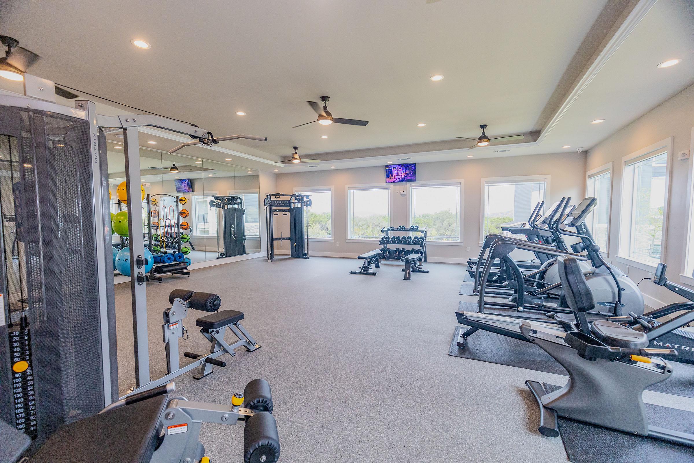 A well-equipped fitness room featuring various gym equipment, including weight machines, free weights, and cardio machines. Large windows allow natural light, and there are ceiling fans for ventilation. The space is designed for both strength training and cardiovascular workouts.