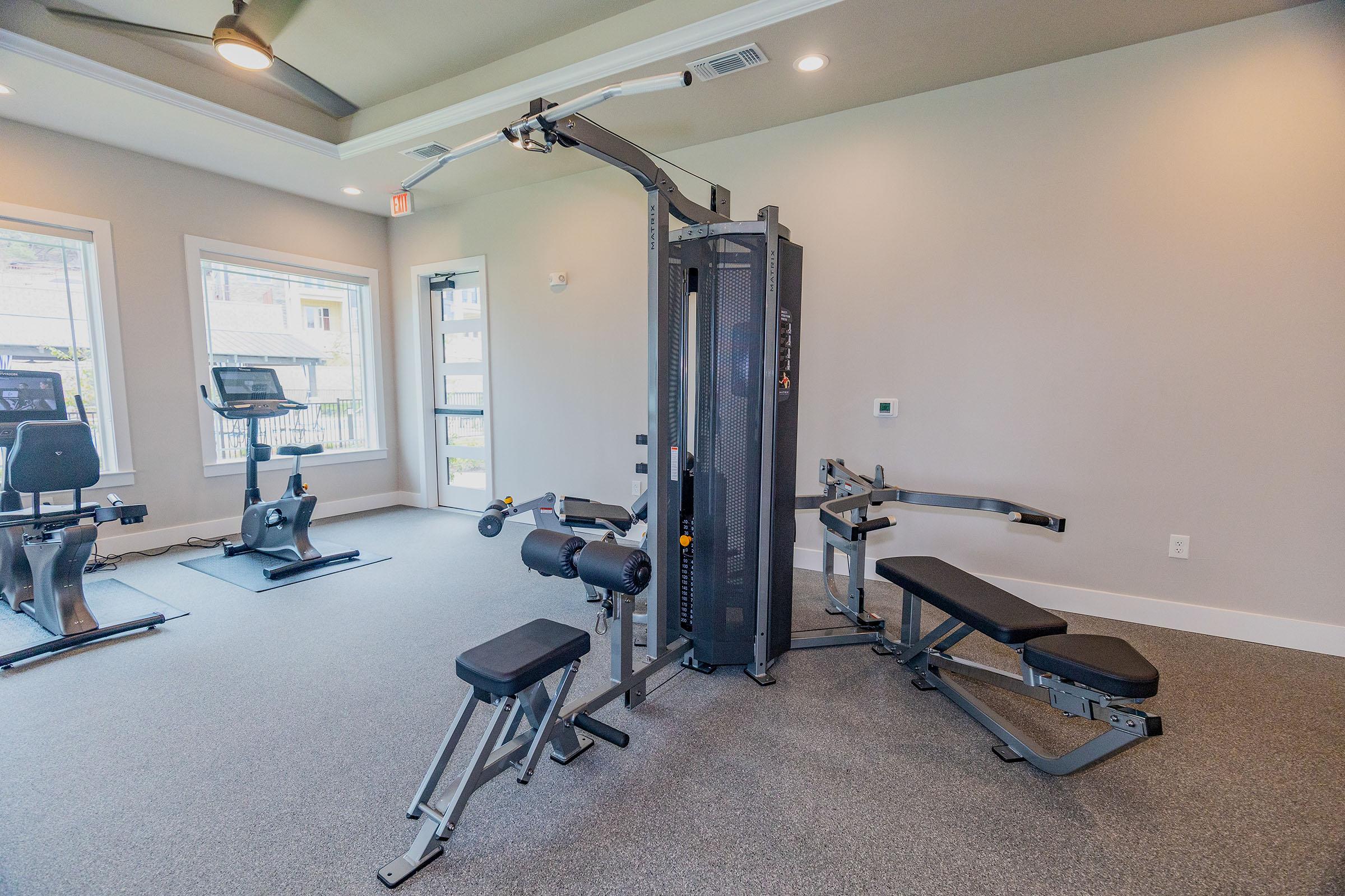 A fitness room featuring a multi-purpose gym machine with a cable system, adjustable seating, and leg extension. Additional exercise equipment is visible, including stationary bikes and treadmills, all situated on gray carpeted flooring and illuminated by natural light from large windows.