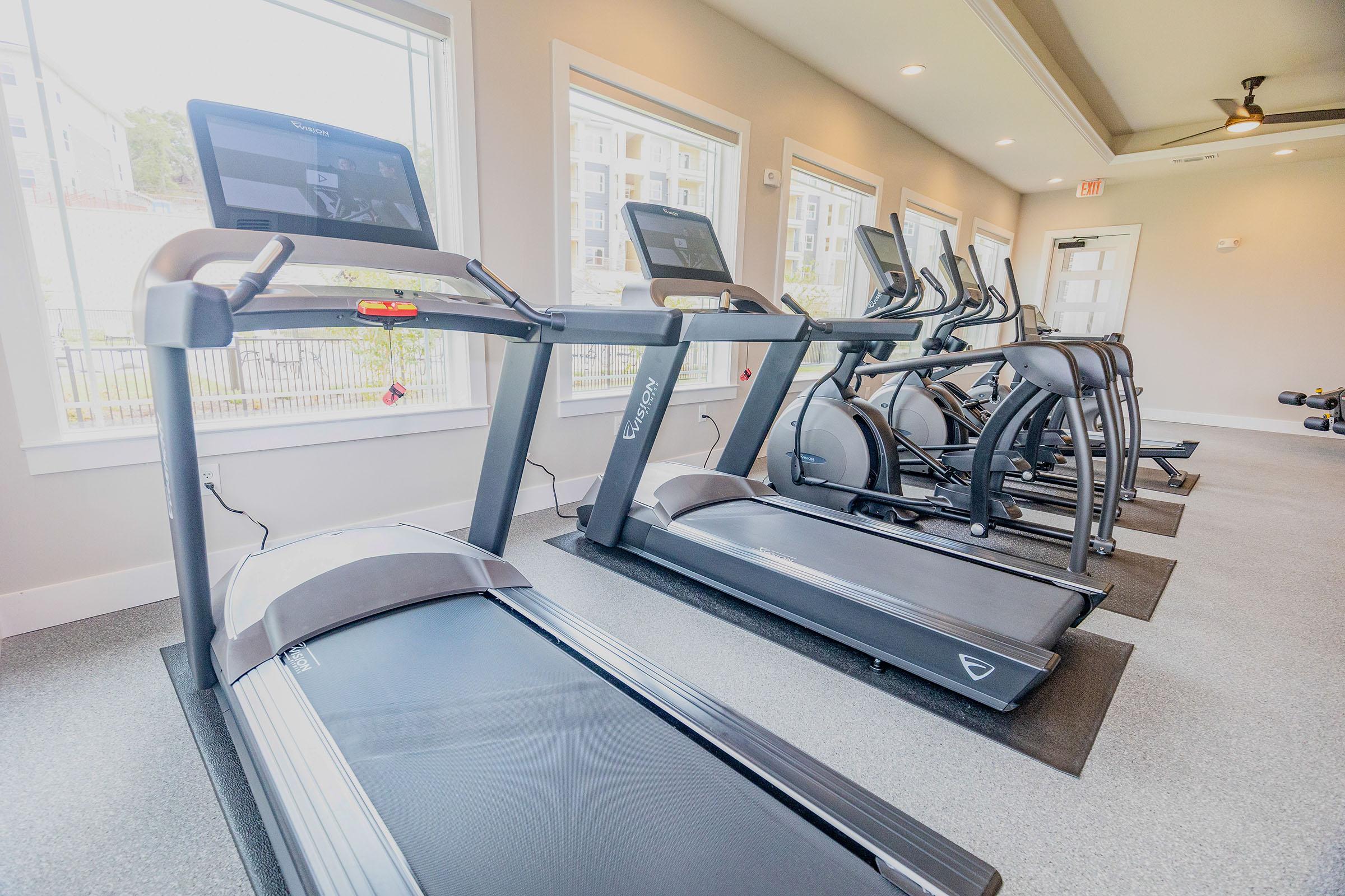 A modern fitness center featuring several exercise machines, including treadmills and elliptical trainers. Large windows provide natural light, and the flooring is carpeted. Screens are mounted on the machines for entertainment or tracking workouts. The space is designed for an active lifestyle.