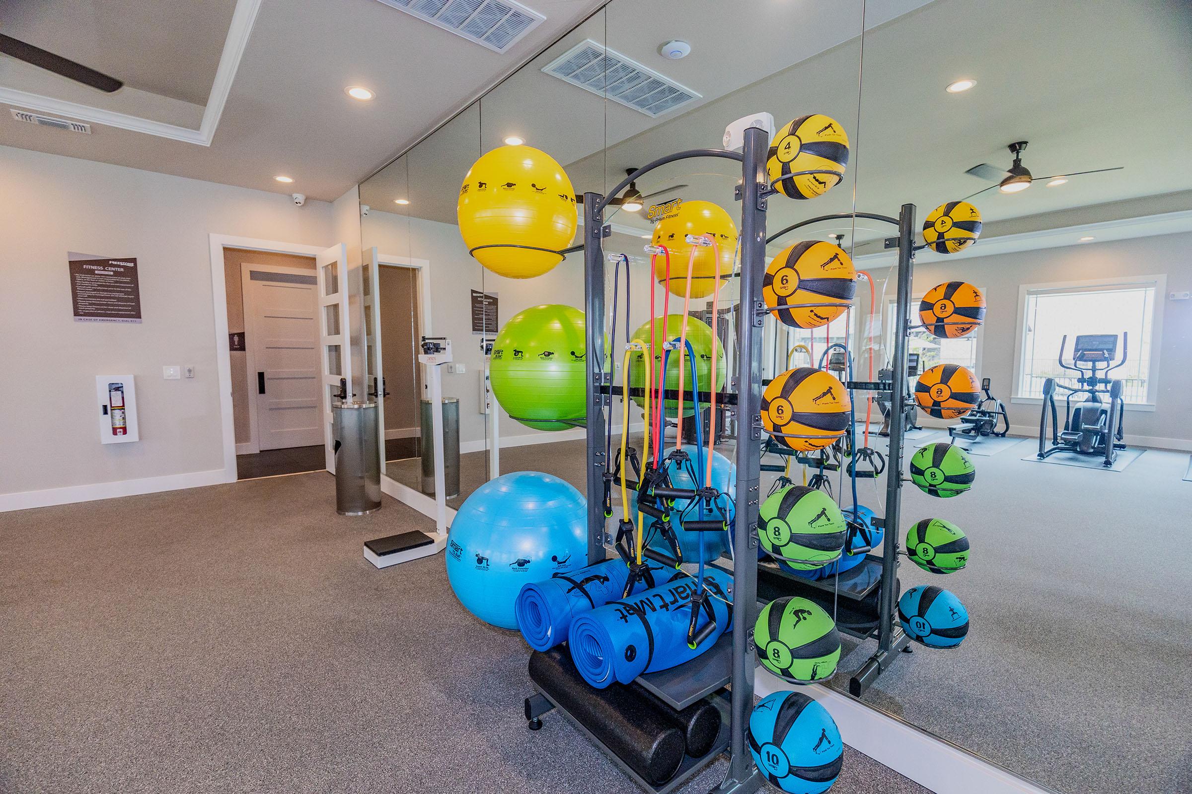 A fitness room featuring various exercise equipment, including colorful medicine balls and stability balls neatly arranged on a rack. A mirror reflects the equipment, and there are exercise mats at the base. The room is well-lit and has a modern design with smooth gray flooring.