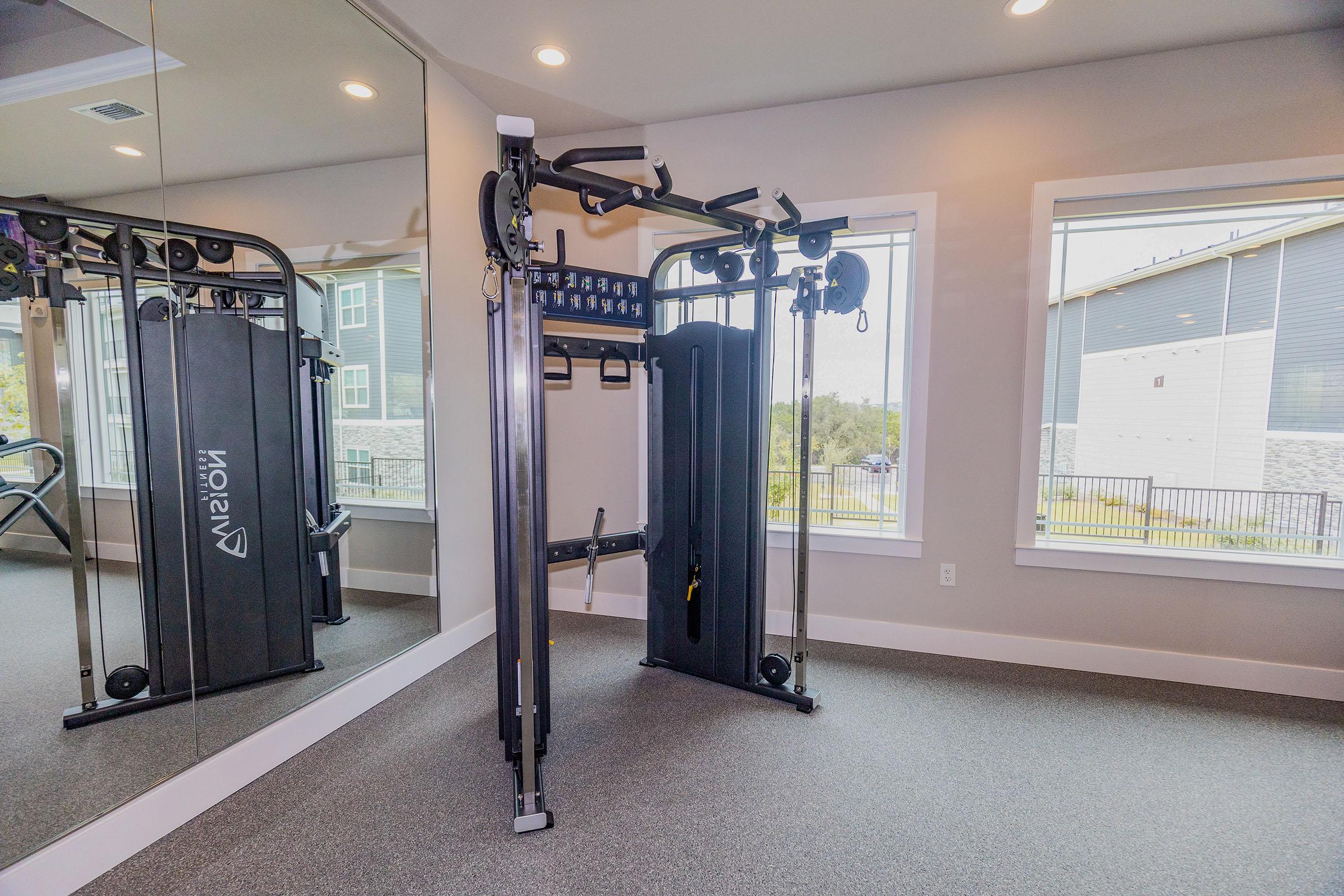 A modern gym space featuring a multifunctional cable machine with various pulley attachments. Mirrored walls reflect the setup, and large windows provide natural light. The flooring is a dark, padded surface suitable for fitness activities. The environment appears clean and well-maintained.