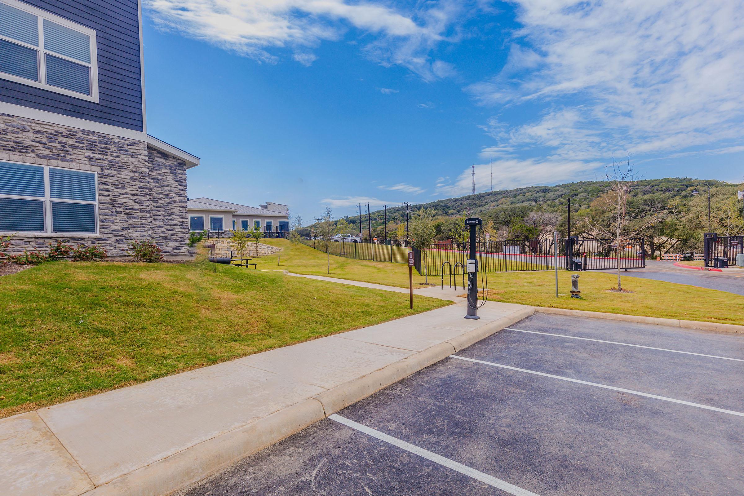 A grassy area next to a parking lot with a bike rack and lamp post, bordered by a stone building on the left. In the background, there's a fence and hills under a clear blue sky with some clouds, indicating a sunny day.