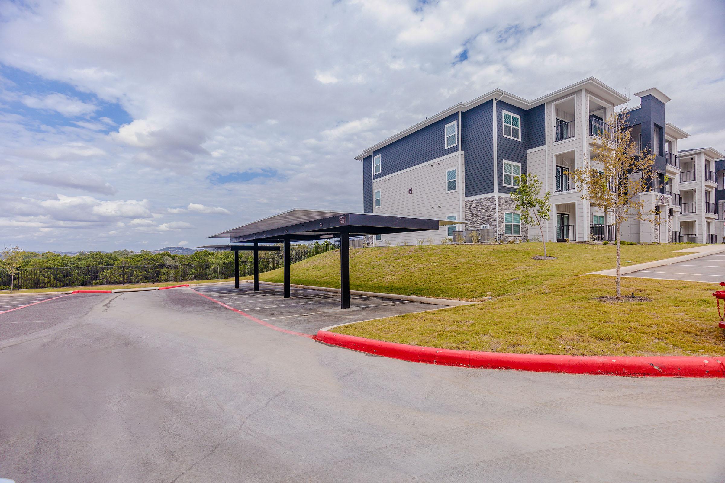 A view of a modern apartment building with gray and blue exterior, set against a cloudy sky. In the foreground, there is a paved parking area with a canopy and a hill covered in greenery. The scene includes well-maintained landscaping and ample space for parking.