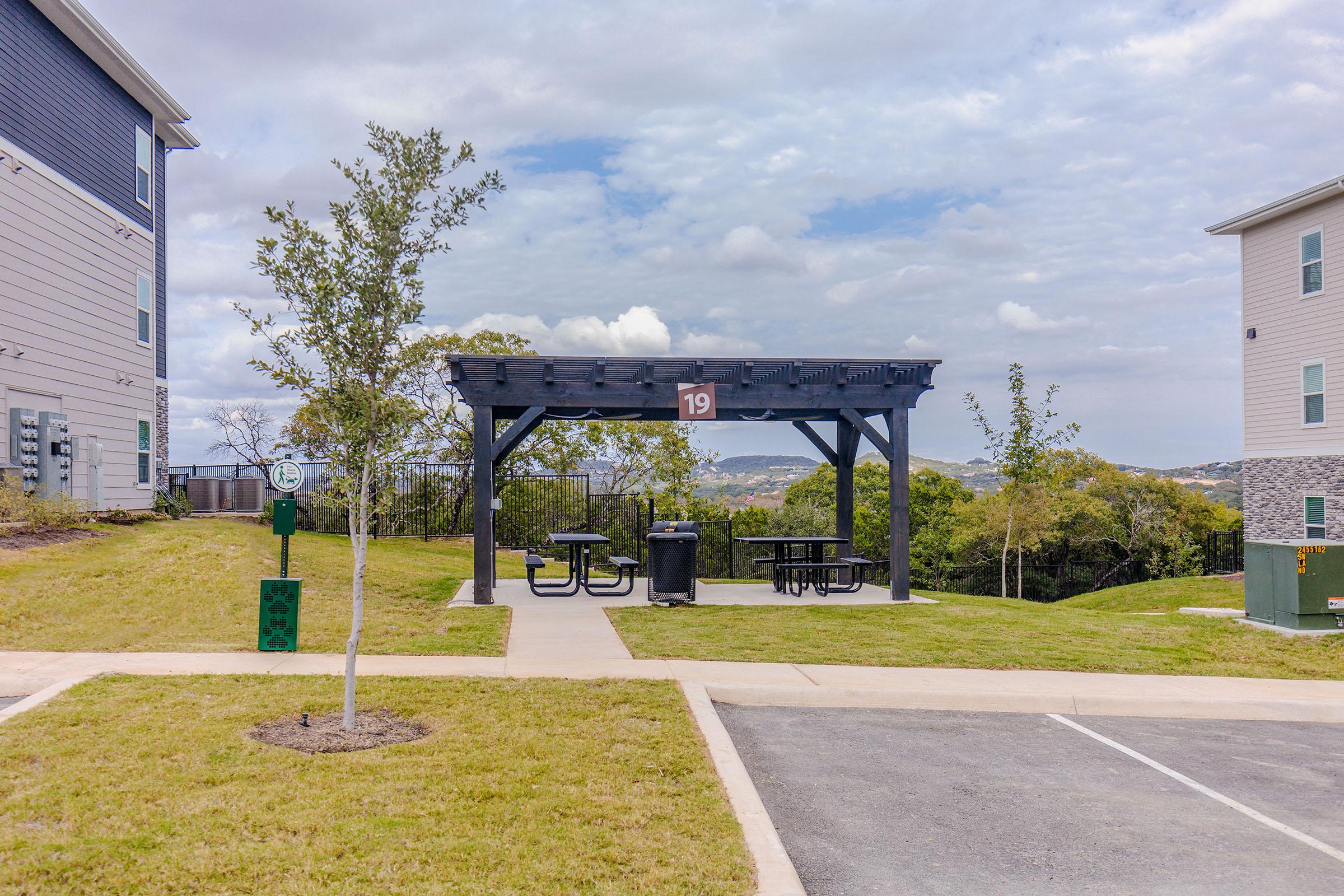 A pavilion with picnic tables and a trash can, marked with the number 19, surrounded by grass and small trees. In the background, there are residential buildings and a cloudy sky with hills visible. The area appears to be a communal outdoor space for relaxation or gatherings.