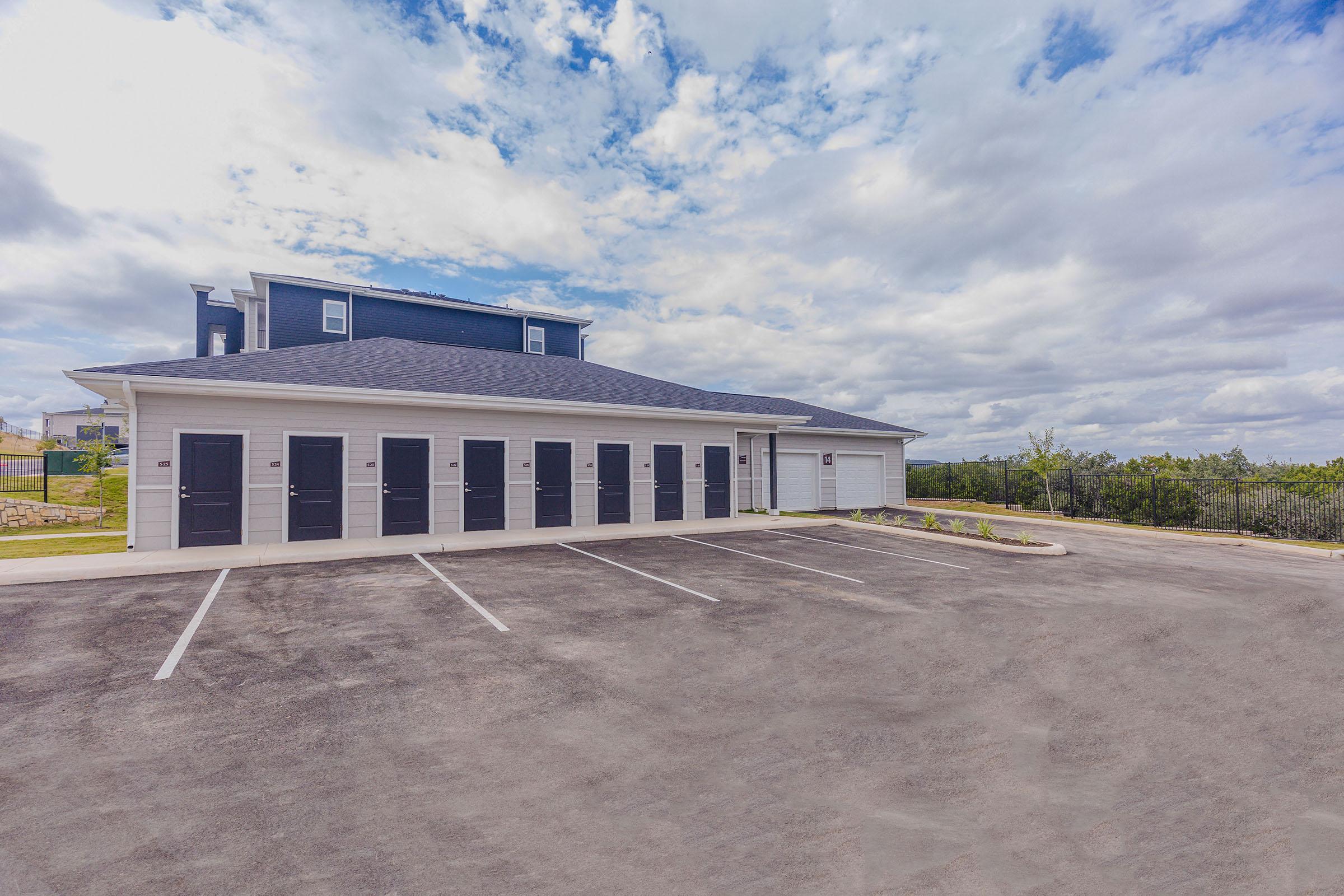 A low two-story building with dark doors lining the front, set against a backdrop of partly cloudy skies. The parking lot in front is empty, and greenery can be seen in the distance, suggesting a rural or suburban area. The structure appears well-maintained and modern.