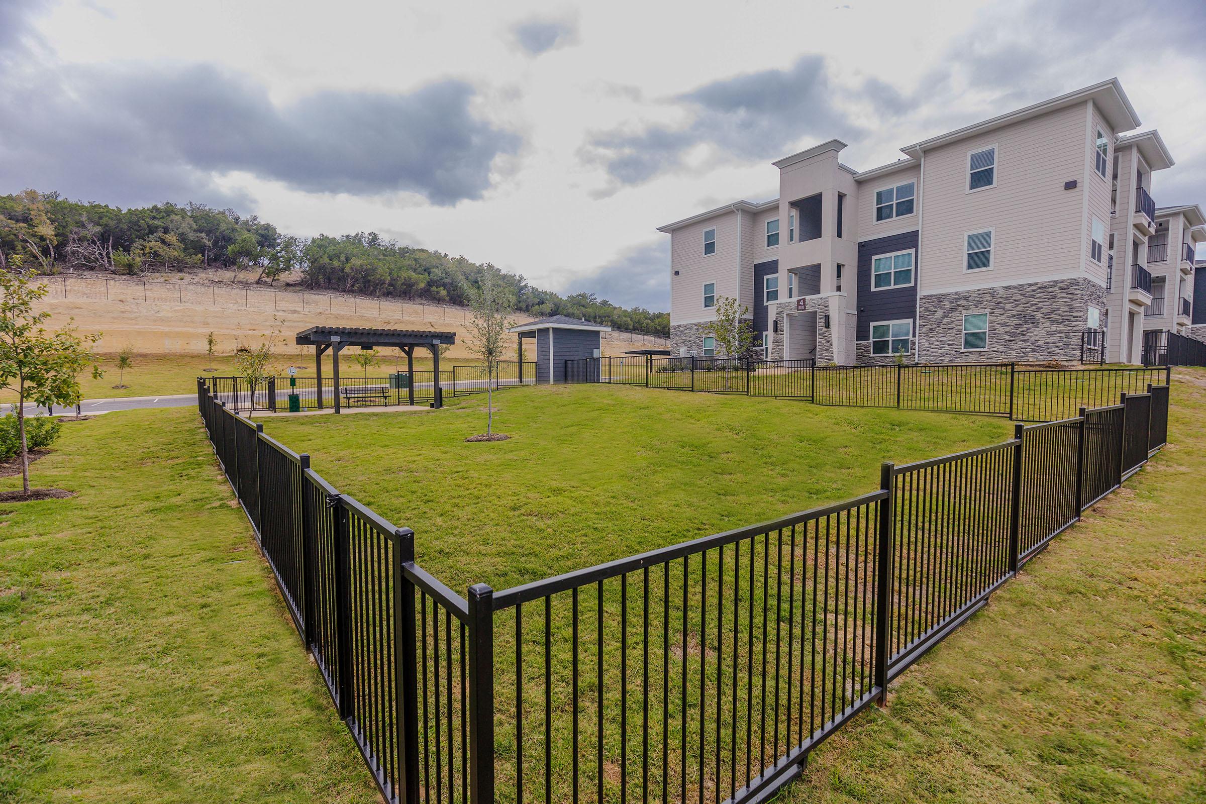 A spacious grassy area enclosed by a black metal fence, featuring small trees and a picnic shelter. In the background, a modern multi-story apartment building is visible, with hills and a cloudy sky behind it. The scene conveys a peaceful residential environment.