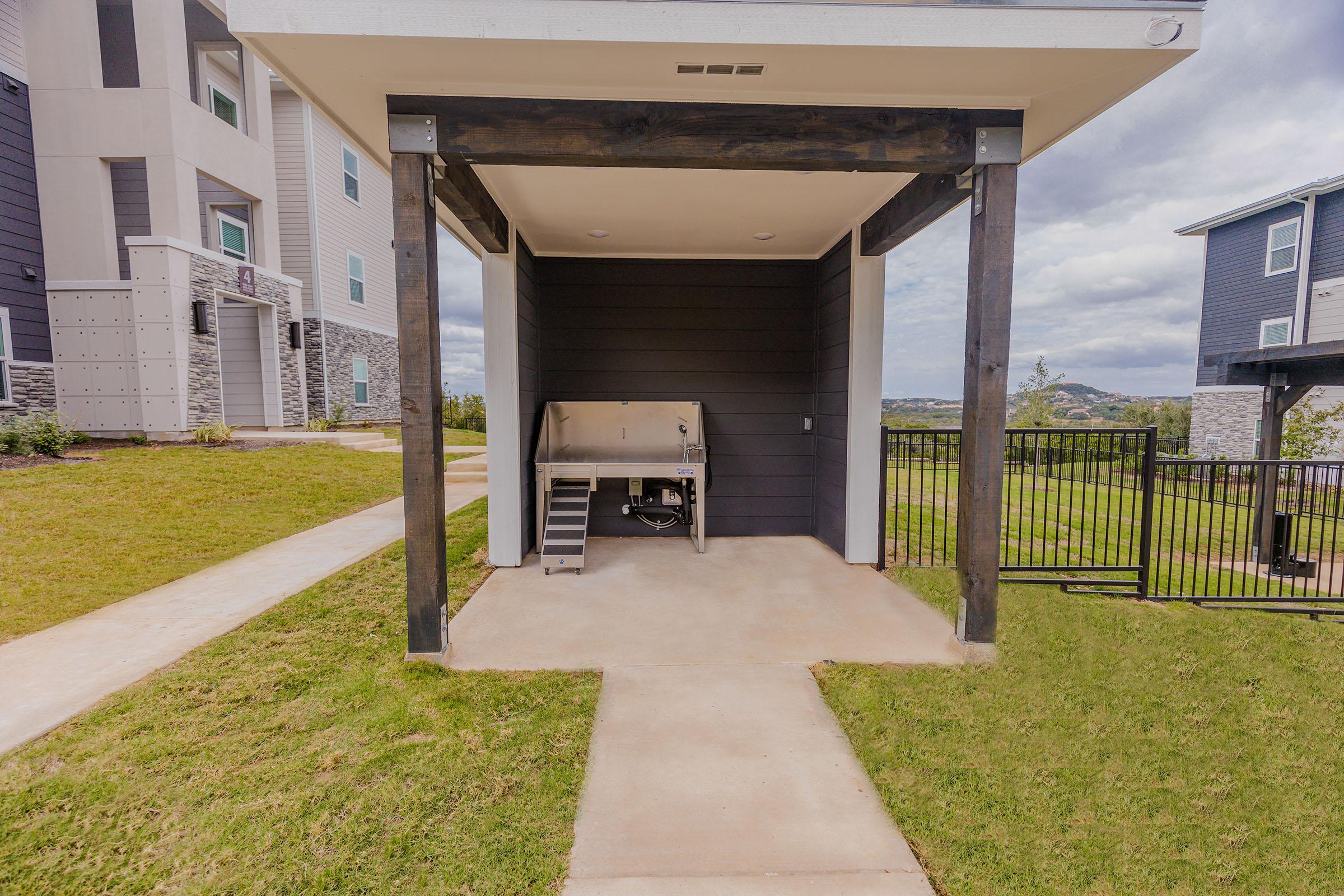 A covered outdoor grilling area featuring a stainless steel grill, situated next to a walkway and surrounded by a grassy yard. The area is part of a modern residential complex with nearby buildings visible in the background. The scene is set under a partly cloudy sky.