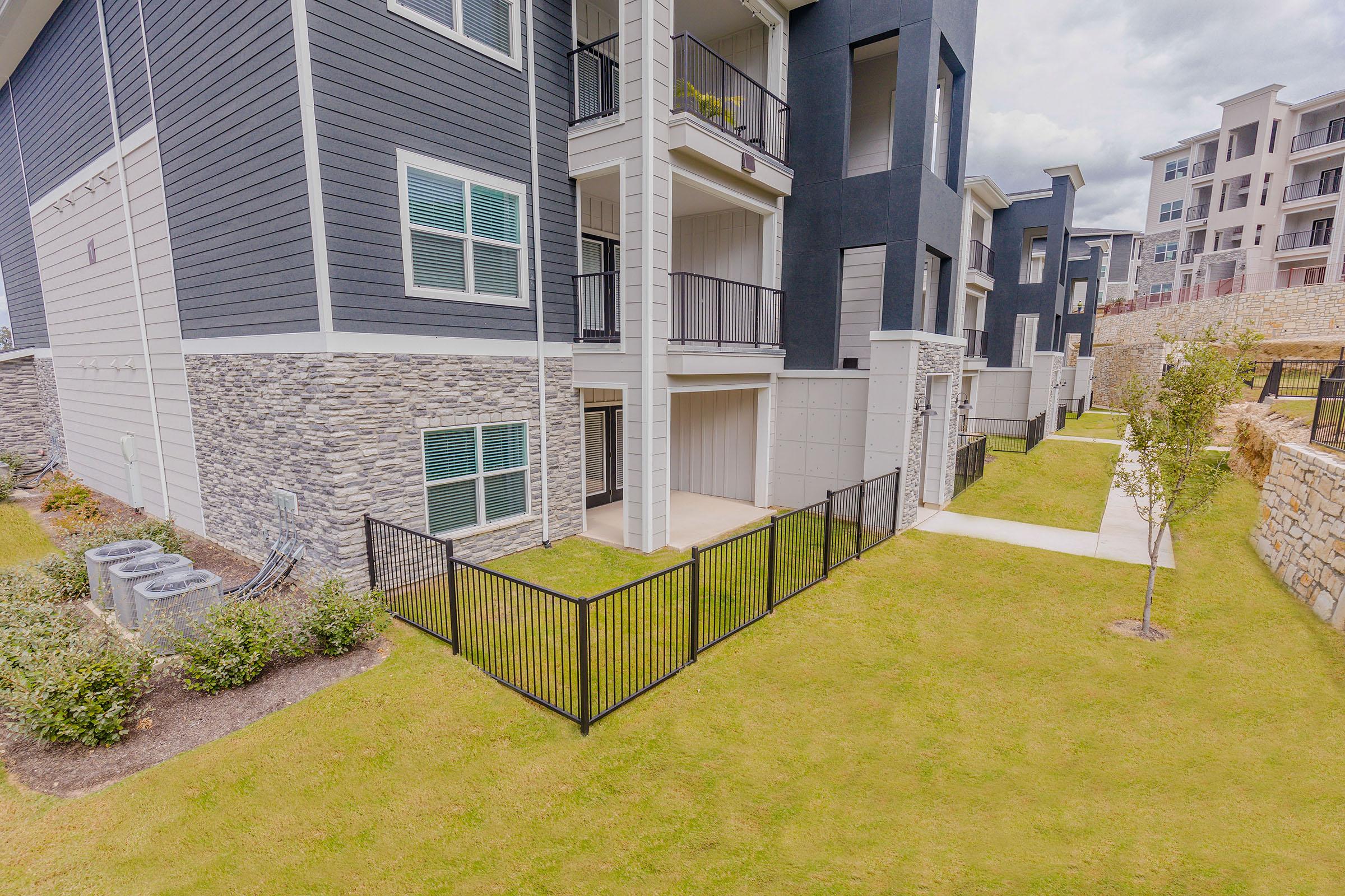 View of a modern apartment complex featuring stacked units with a mix of gray siding and stone accents. The ground level includes a small fenced yard, and well-manicured grass surrounds the building. The image captures a portion of the complex, highlighting pathways and landscaping.