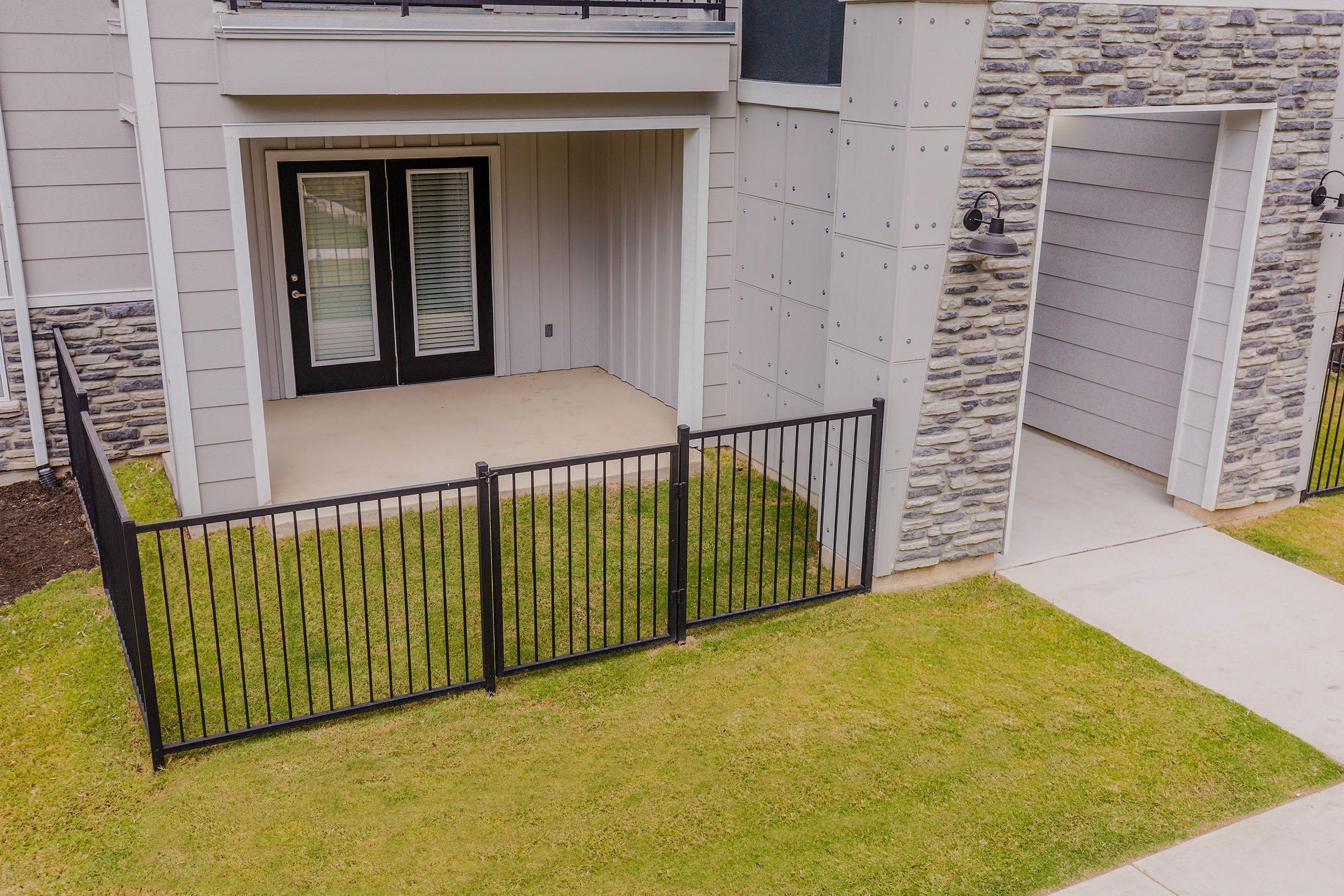 A view of a minimal outdoor space beneath an apartment balcony, featuring a small, fenced grass area. The space includes double glass doors leading to the interior and a stone wall beside a pathway.